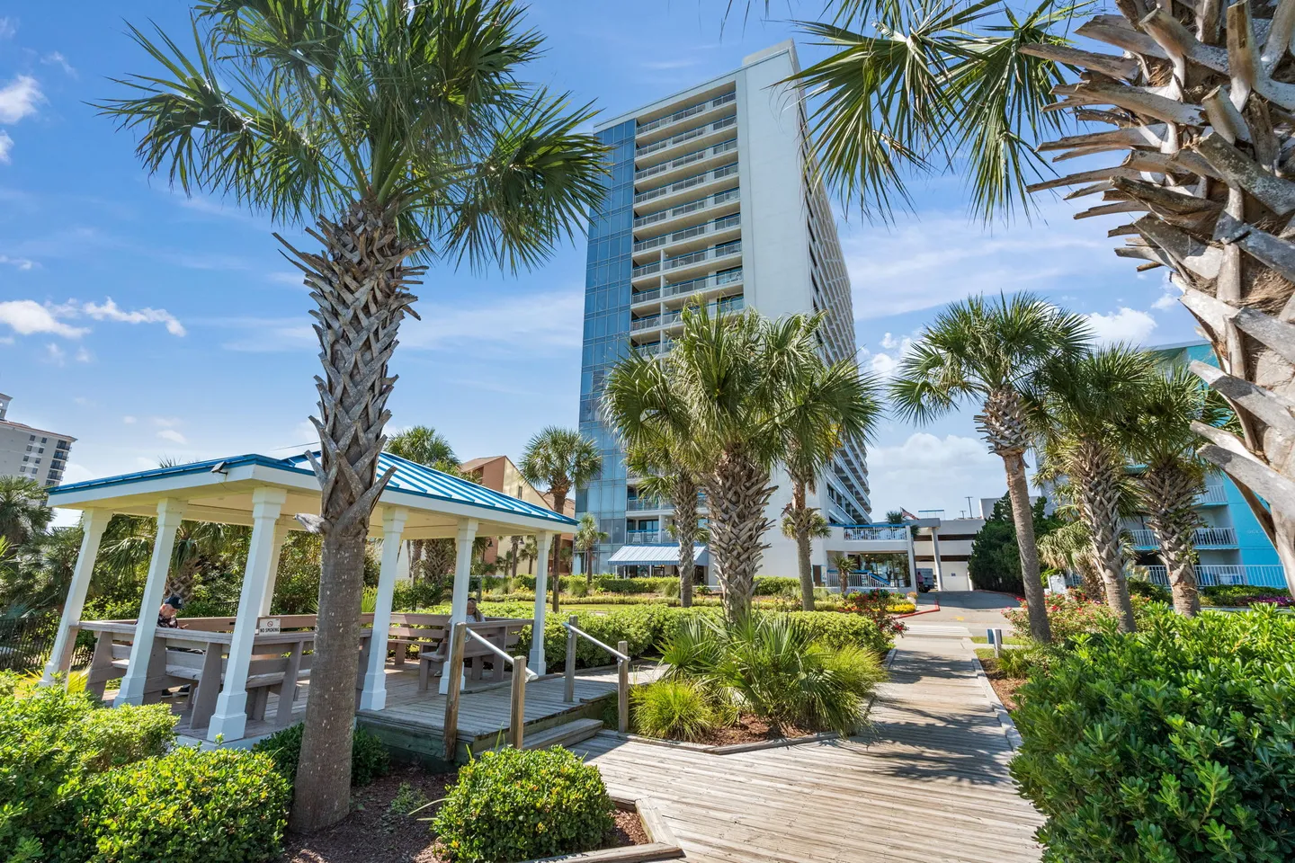 Outdoor area featuring a wooden boardwalk, covered picnic gazebo, palm trees, and a multistory condominium building under a sunny sky.