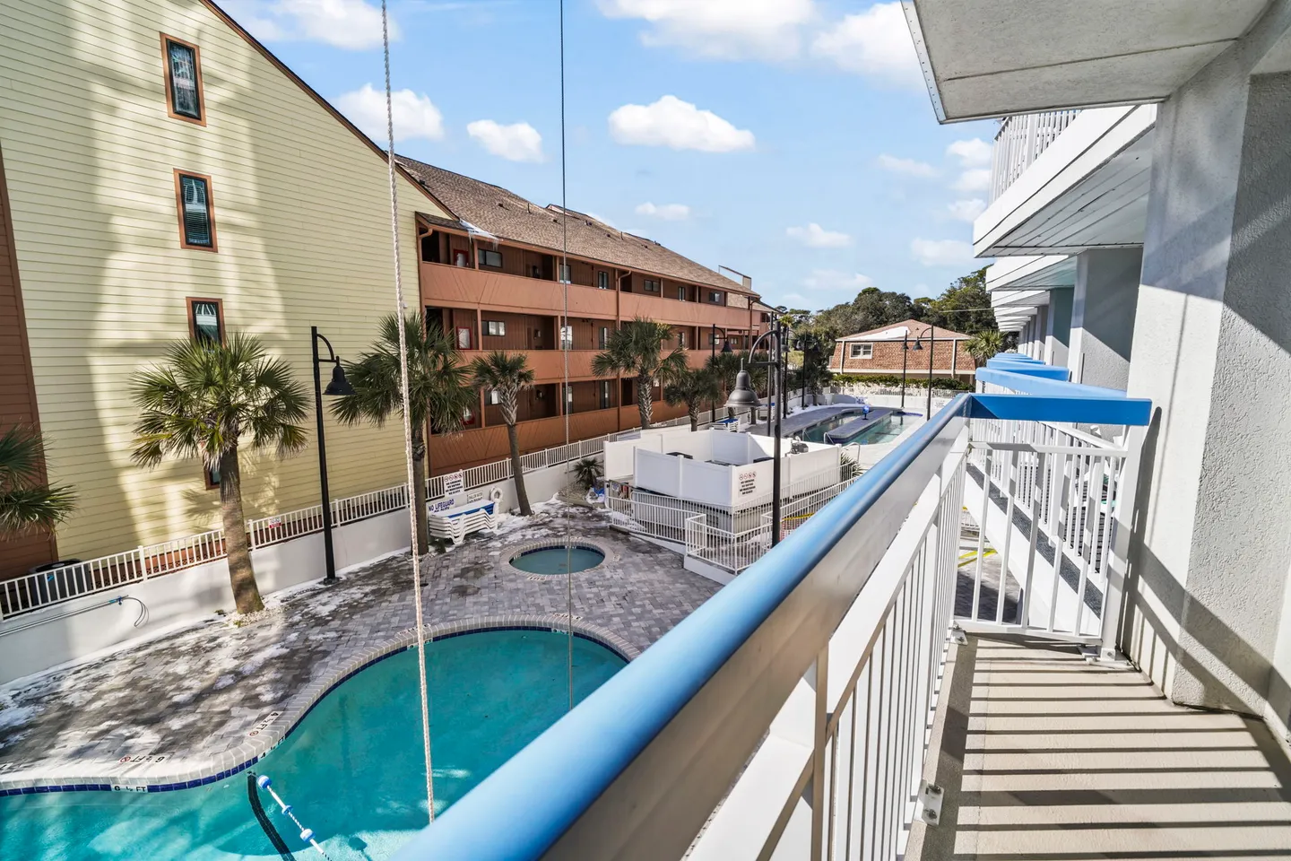 Balcony view overlooking a fenced courtyard with swimming pools and palm trees between multi-story residential buildings.