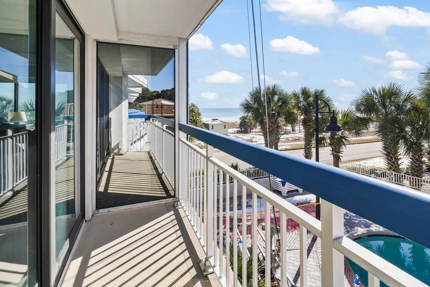 Balcony with white and blue railing overlooking palm trees, a swimming pool, and the ocean under a blue sky.