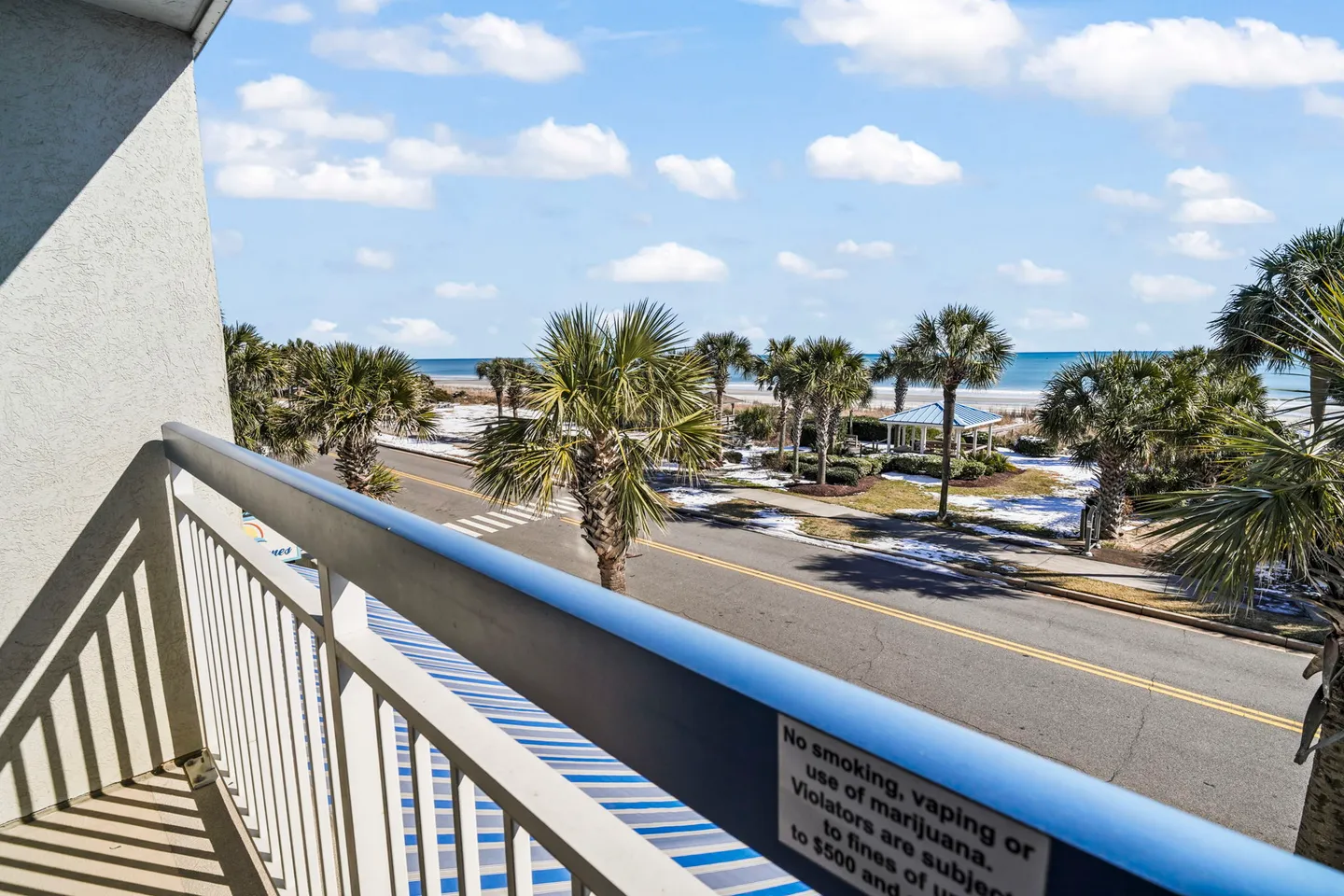Balcony view overlooking a street with palm trees, a beach, and the ocean under a partly cloudy sky.
