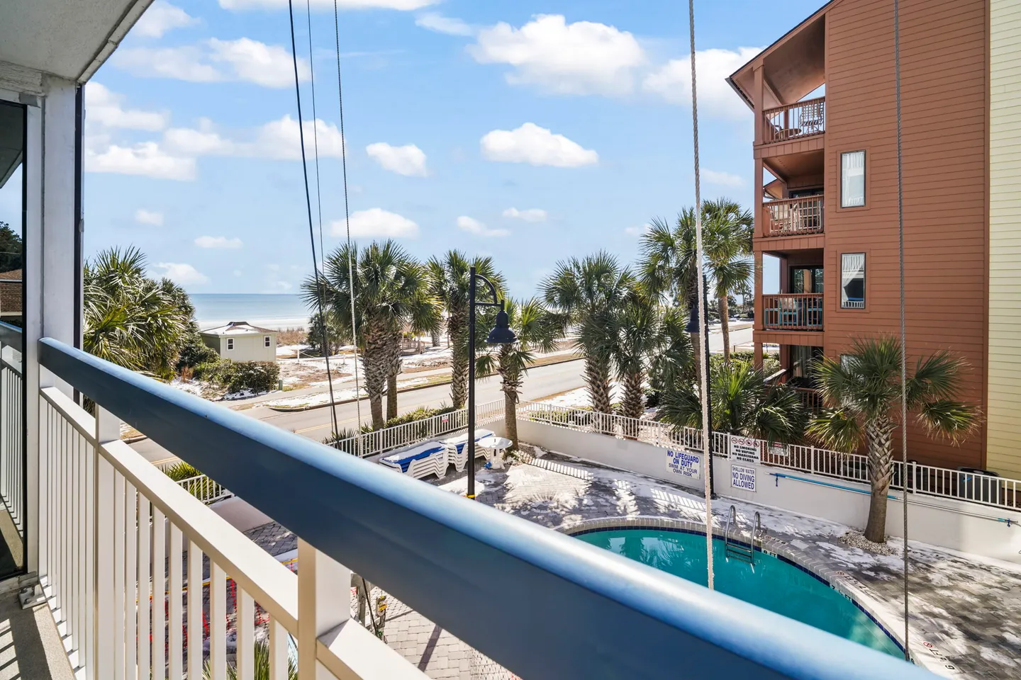 Balcony view overlooking a swimming pool patio, palm trees, and the beach and ocean in the distance.