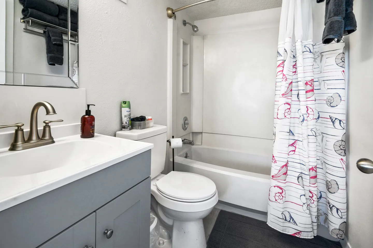 Bathroom with a grey vanity, a white toilet, and a bathtub with a shower curtain patterned with seashells.