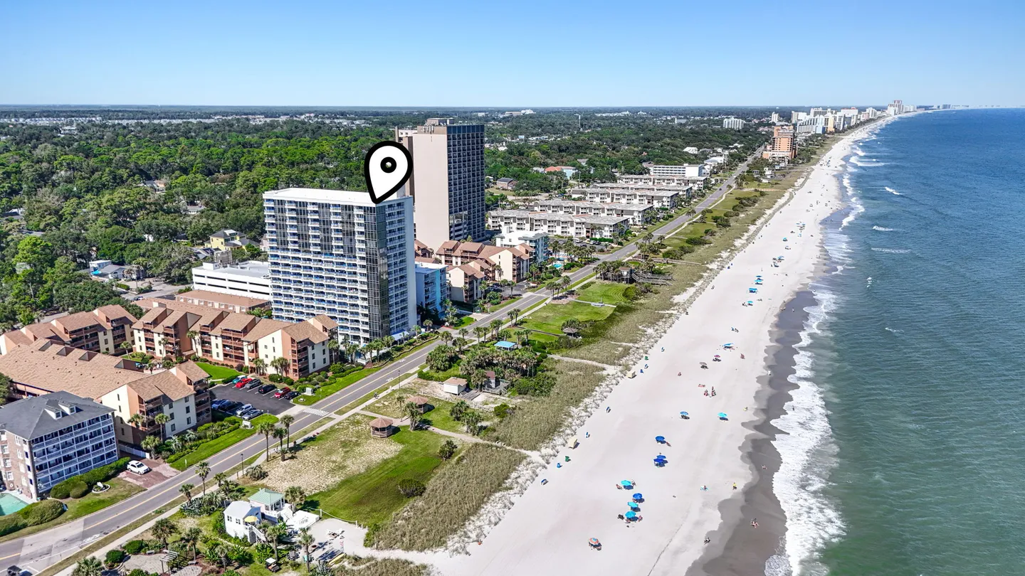 Aerial view of a multistory beachfront condominium building marked by a location pin, alongside a sandy coastline and the ocean.