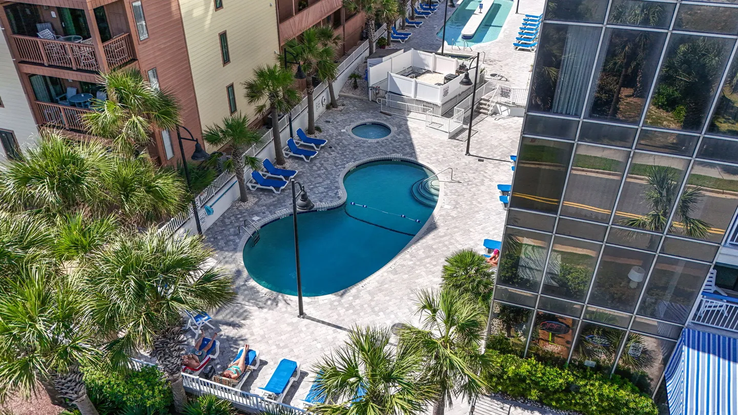High-angle view of an outdoor community swimming pool, hot tub, and sun deck with lounge chairs surrounded by palm trees and multi-story buildings.