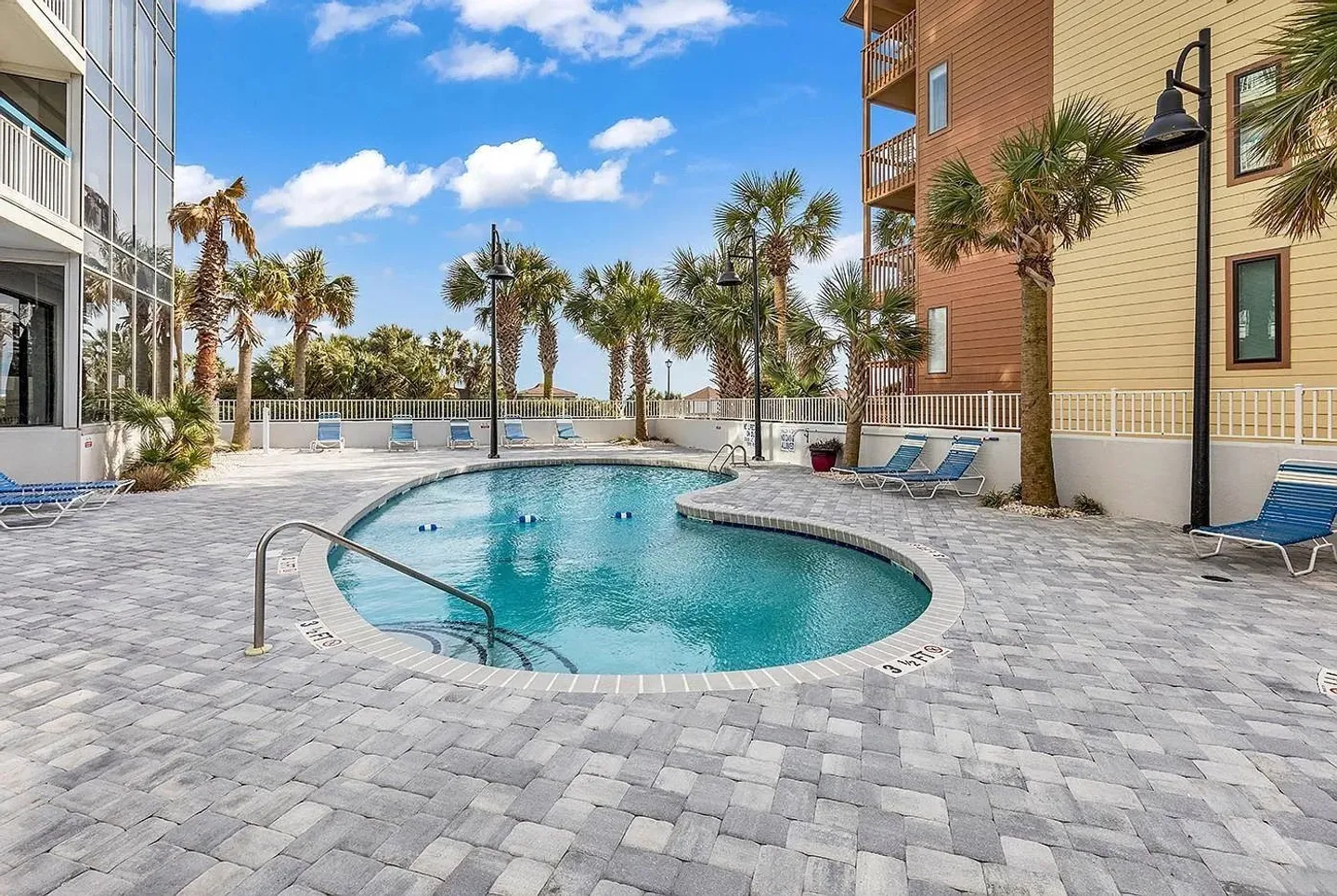 Outdoor swimming pool surrounded by paver patio, lounge chairs, palm trees, and residential buildings.
