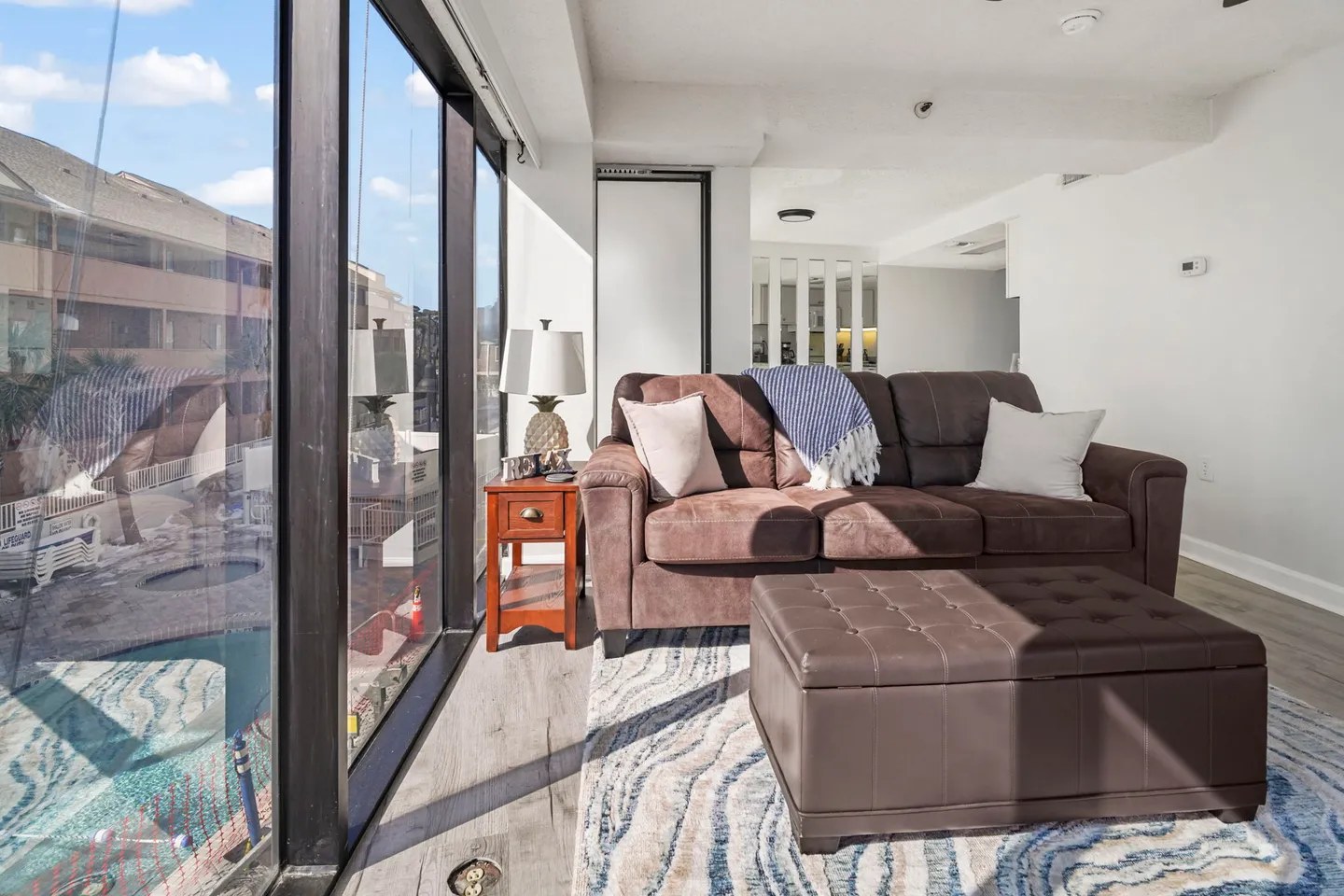 Sunlit living area with a brown couch and large floor-to-ceiling windows looking out toward an outdoor pool area.