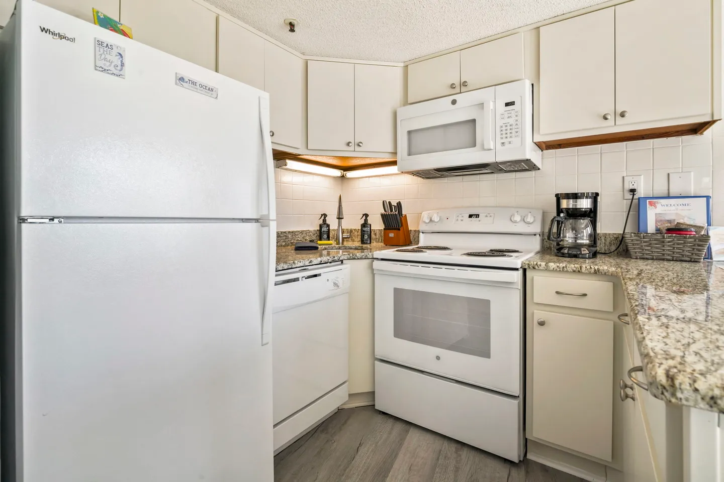 Kitchen corner showing white appliances including a refrigerator, dishwasher, stove, and microwave alongside speckled countertops and light-colored cabinets.