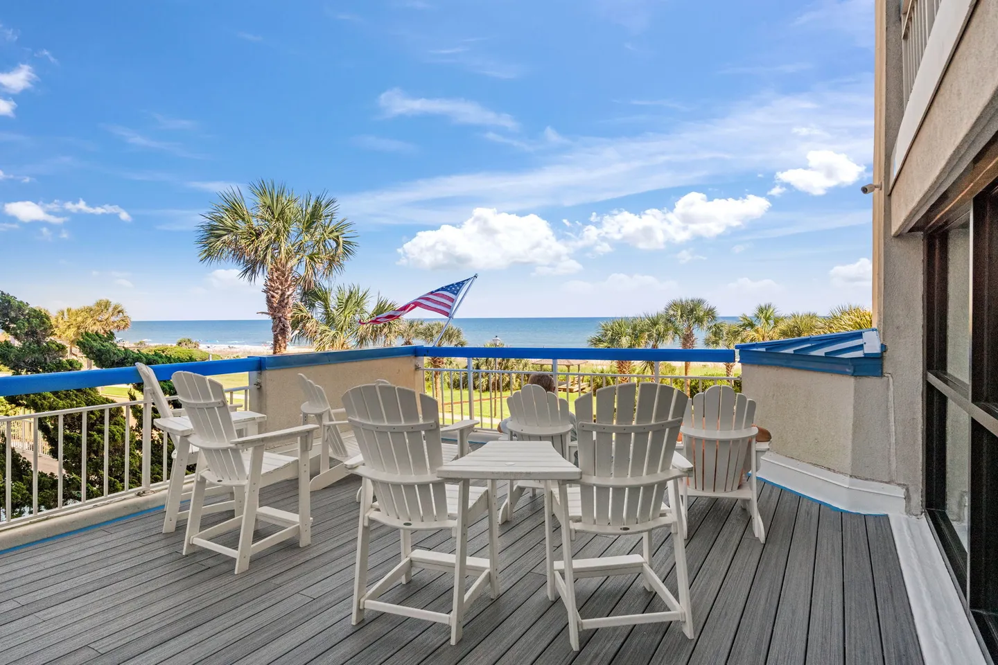 Outdoor balcony with grey decking, white chairs, and an ocean view under a blue sky.