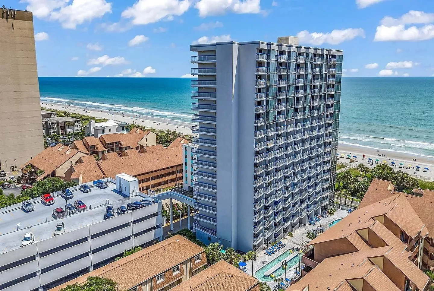 Aerial view of a high-rise oceanfront condominium building next to a parking garage, pool, and sandy beach.