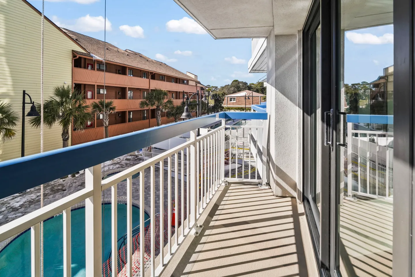 Balcony with white and blue railing overlooking a swimming pool and neighboring apartment buildings under a blue sky.