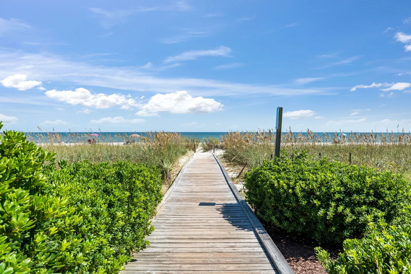A wooden boardwalk path flanked by green bushes leading toward a sandy beach and blue ocean under a partly cloudy sky.