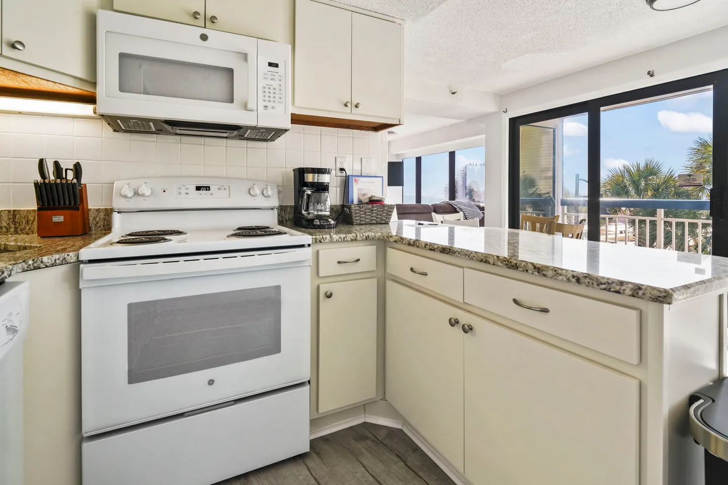 A kitchen with white appliances, speckled countertops, and a view through sliding glass doors to a balcony and palm trees.