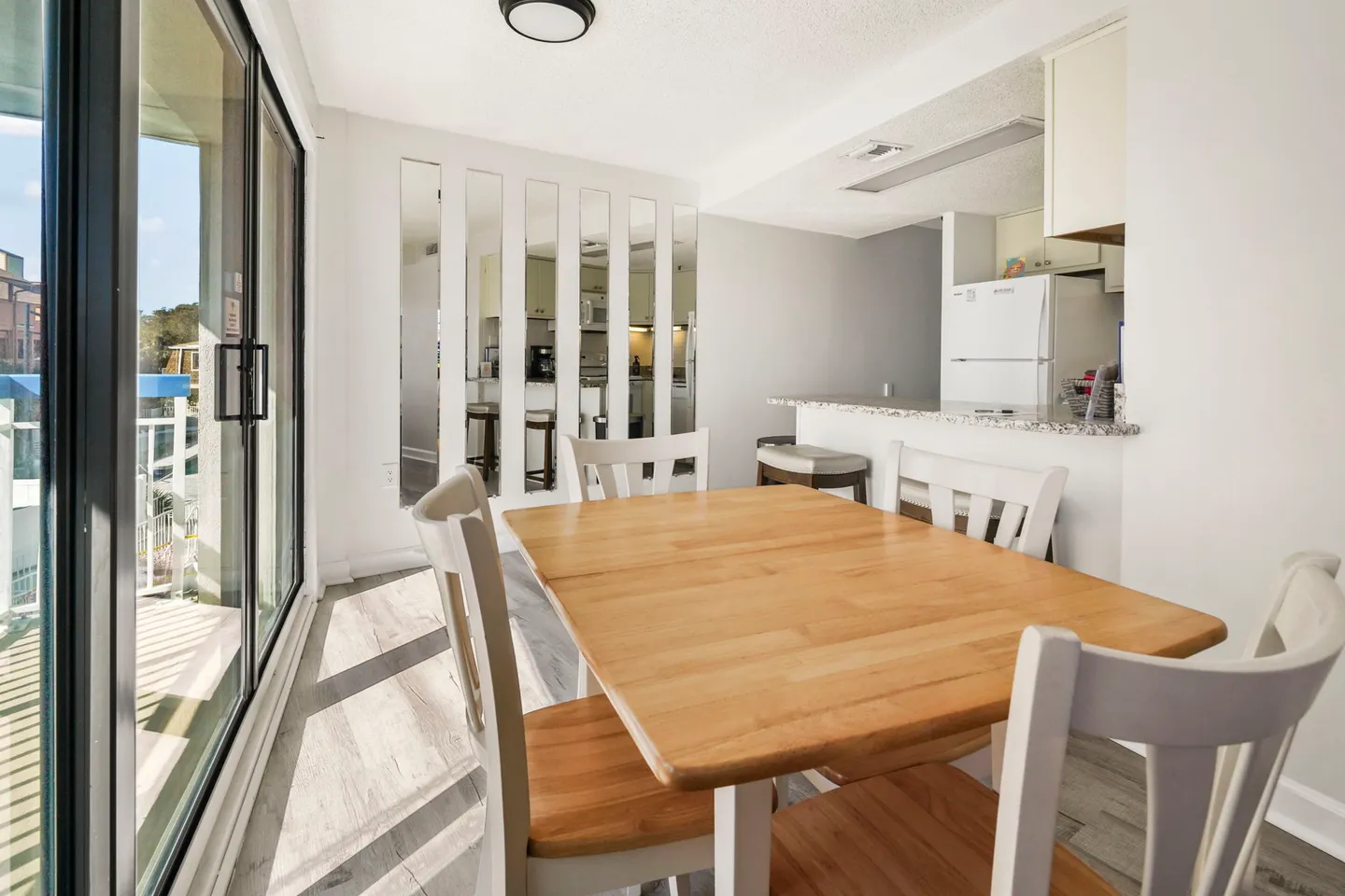 Dining area with a wood-top table, white chairs, sliding glass door leading to a balcony, and a kitchen counter in the background.