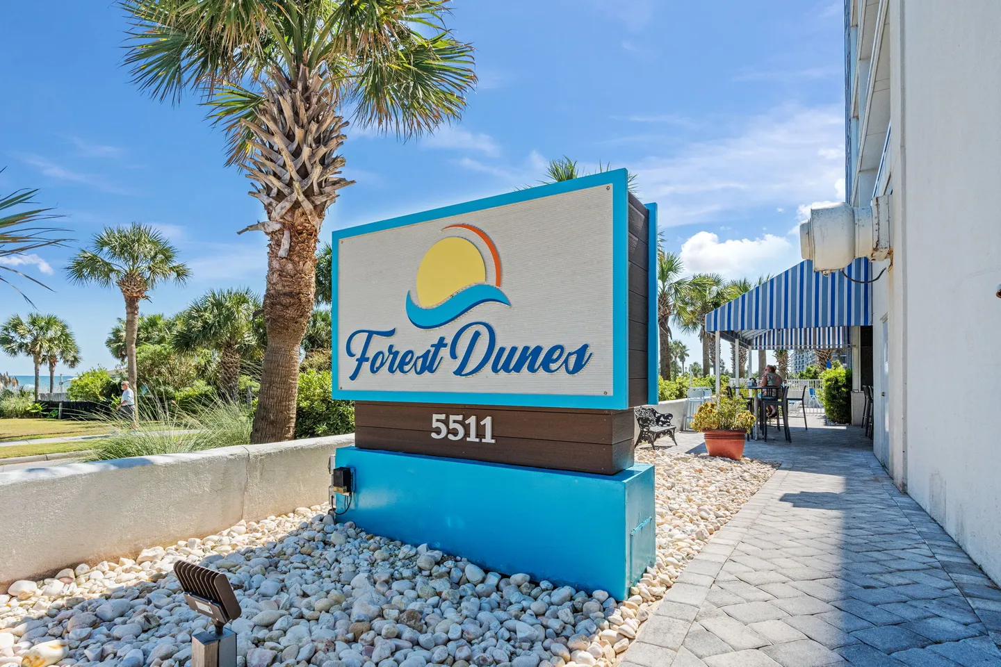 Outdoor property sign for Forest Dunes with a sun logo, palm trees, and a paved walkway under a blue sky.