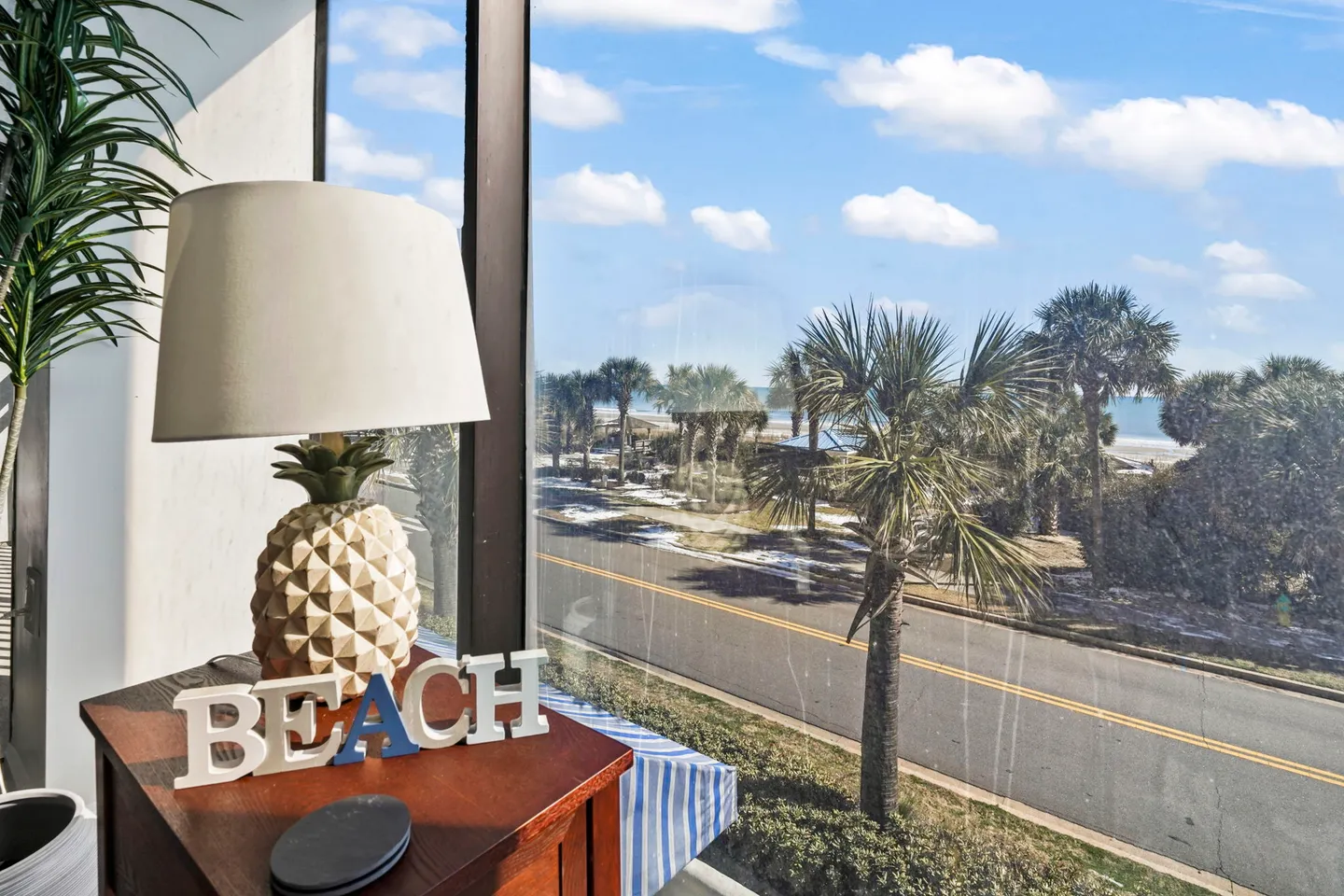 Window view of a road, palm trees, and the ocean beyond, framed by a side table with a pineapple lamp and decorative BEACH letters indoors.