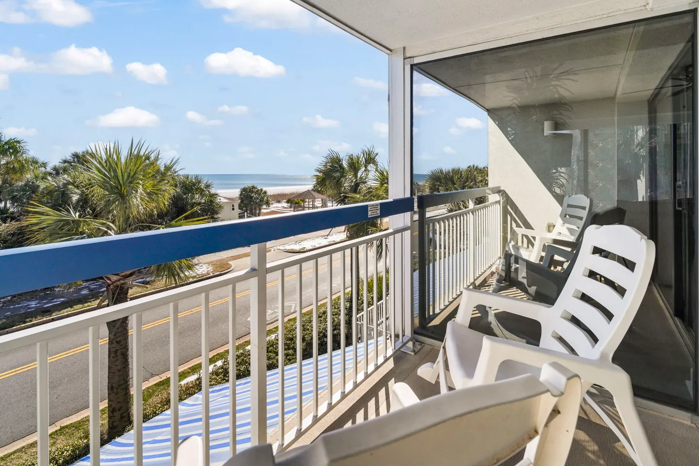 Balcony with white plastic chairs overlooking palm trees, a road, and the ocean beach under a blue sky.