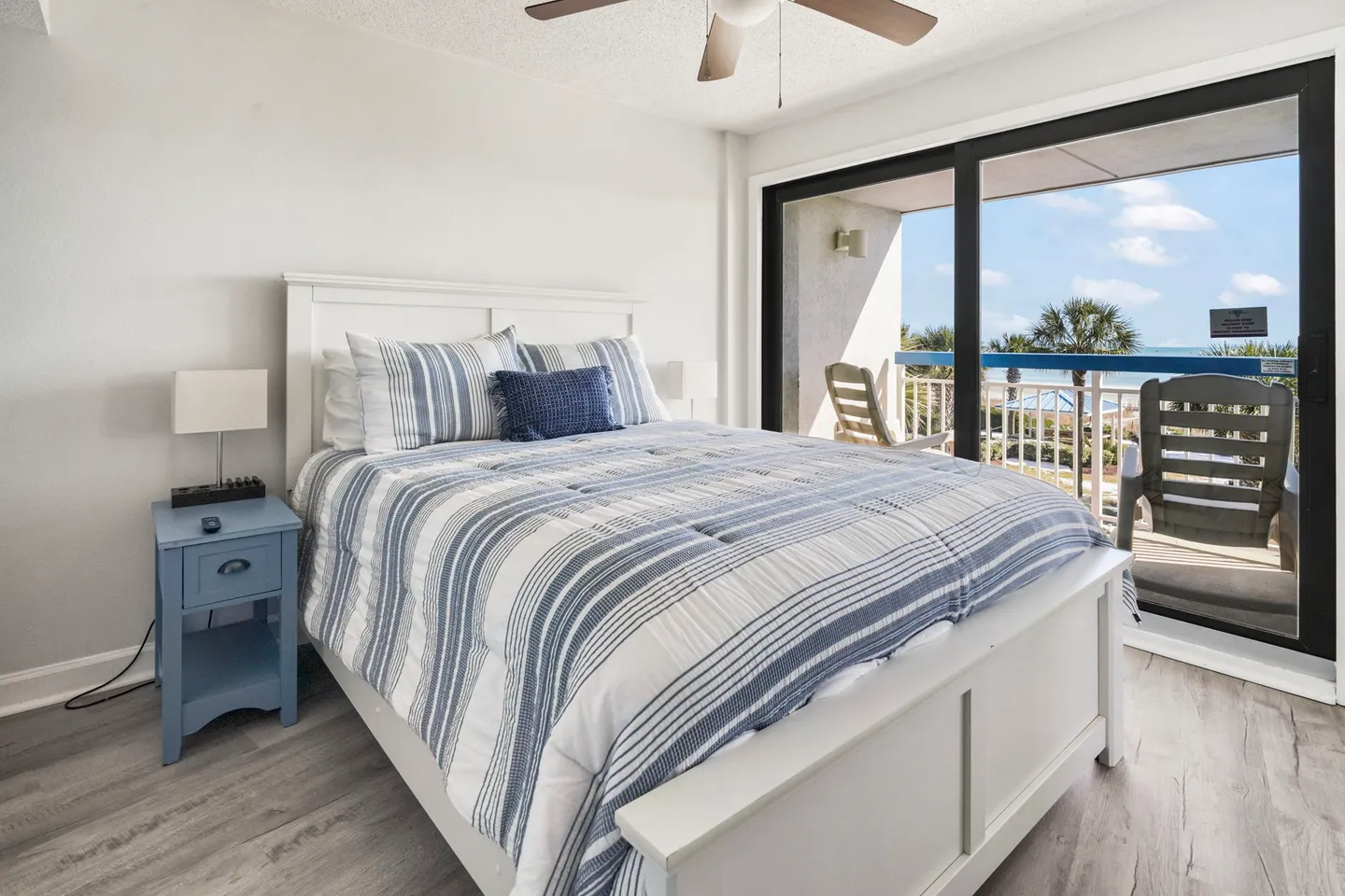 Bedroom with a white panel bed dressed in blue and white striped bedding, next to sliding glass doors opening to a balcony with ocean views.