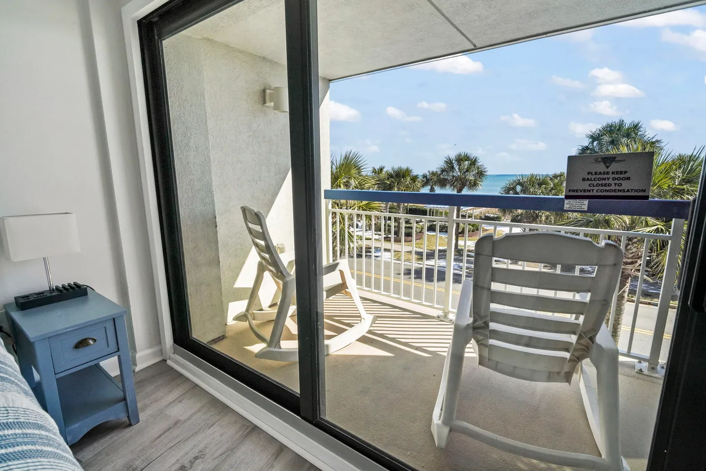 Sliding glass doors opening onto a covered balcony with two white rocking chairs overlooking palm trees and the ocean.