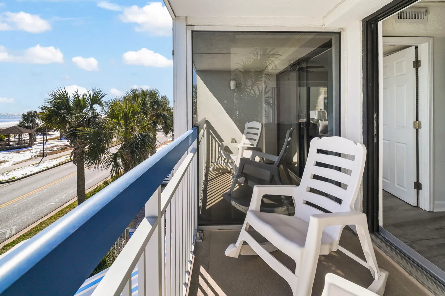 Covered balcony with white rocking chairs overlooking palm trees, a road, and the coast.