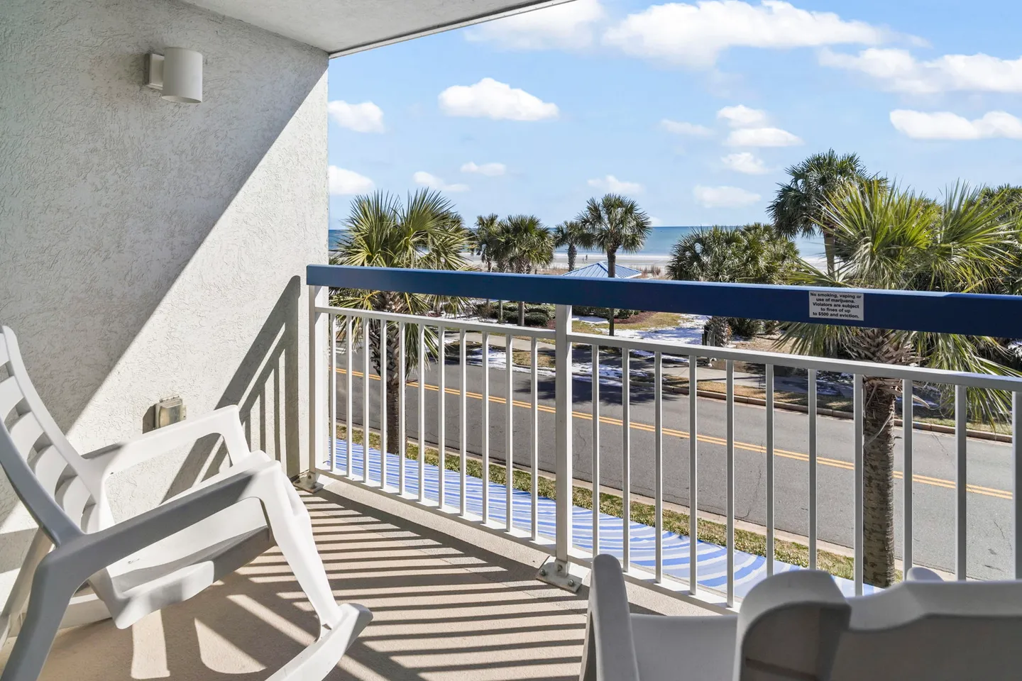 Covered balcony with white rocking chairs overlooking a road, palm trees, and the ocean under a blue sky.