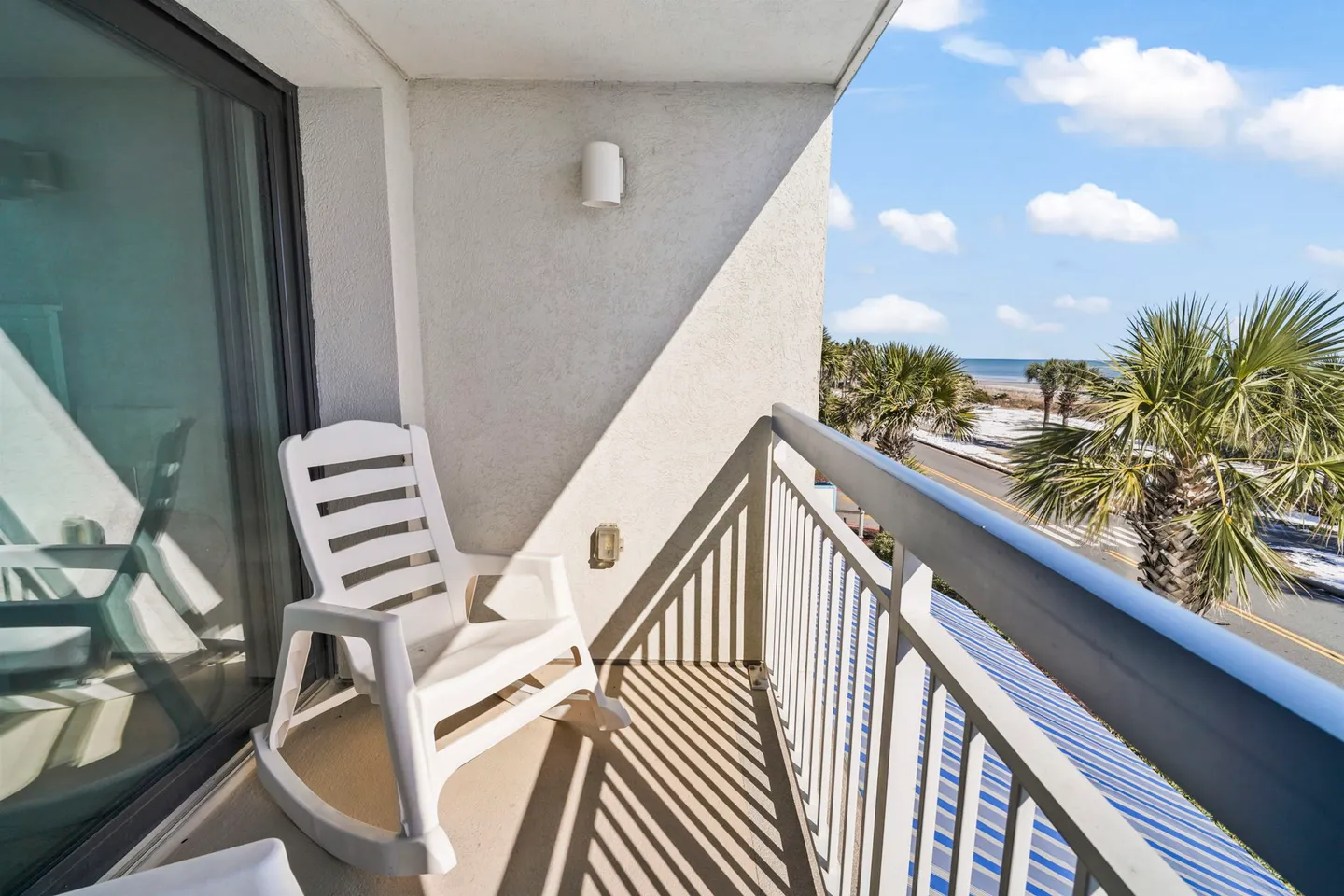 A private outdoor balcony featuring a white rocking chair, railings, and a view of palm trees, a road, and the ocean under a blue sky.