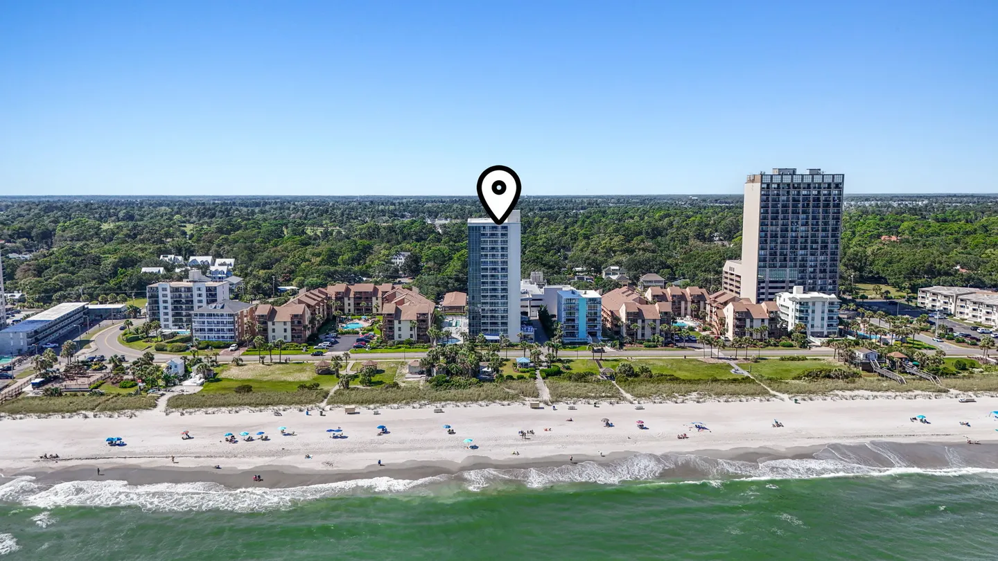 Aerial view of an oceanfront multi-story building marked by a location pin along a sandy beach with breaking ocean waves.