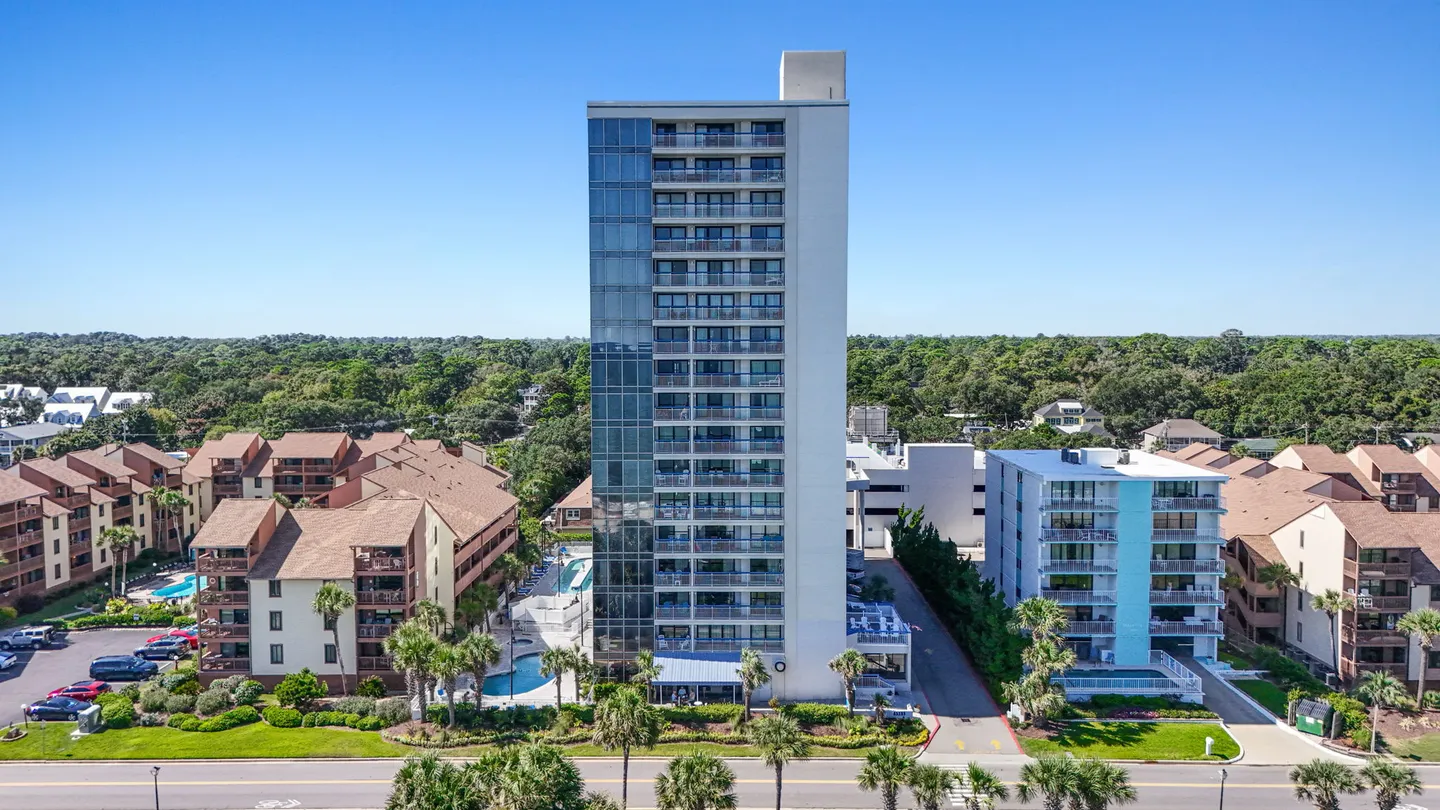 Aerial view of a tall multi-story beachfront condominium building surrounded by neighboring resorts, palm trees, and swimming pools under a clear sky.