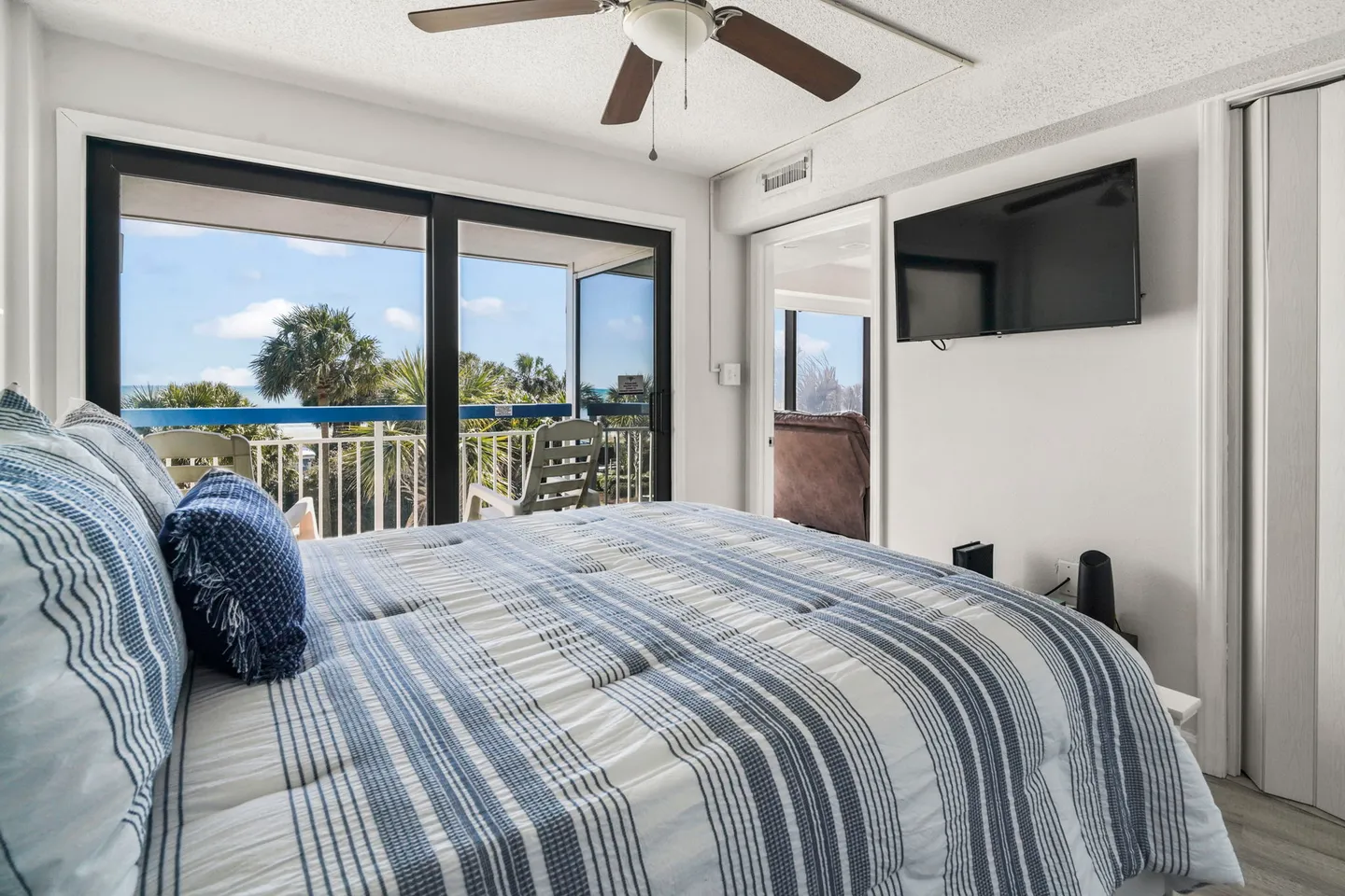 Bedroom with blue striped bedding, ceiling fan, wall-mounted television, and sliding glass doors leading to a balcony.