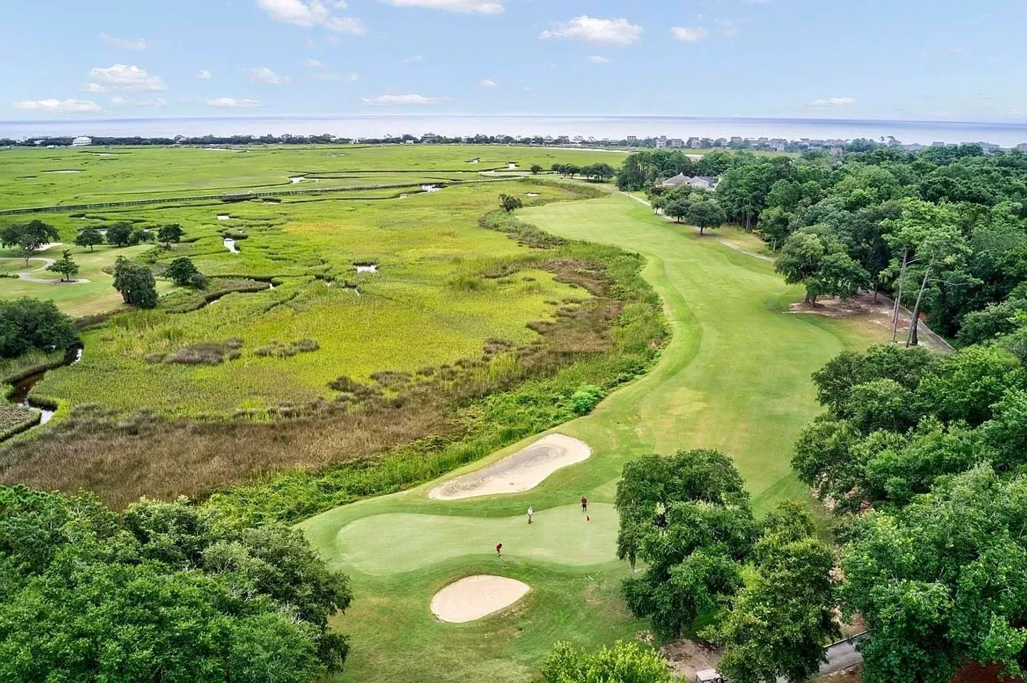 Aerial view of a green golf course bordering a marsh with the ocean visible in the background under a blue sky.
