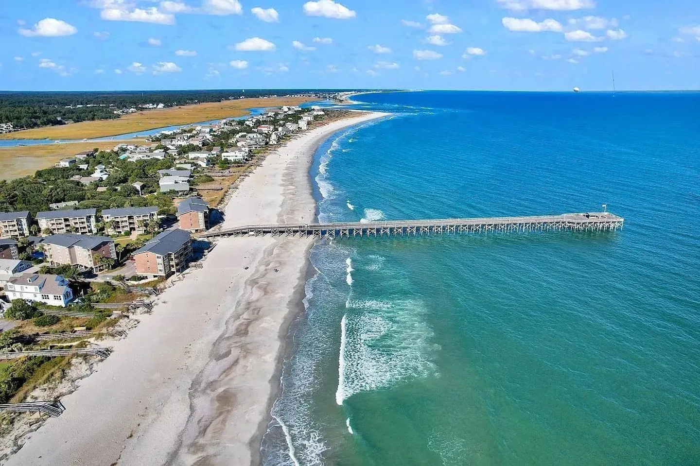 Aerial view of a sandy coastline with ocean waves, beachfront buildings, and a long wooden pier extending into the turquoise water.
