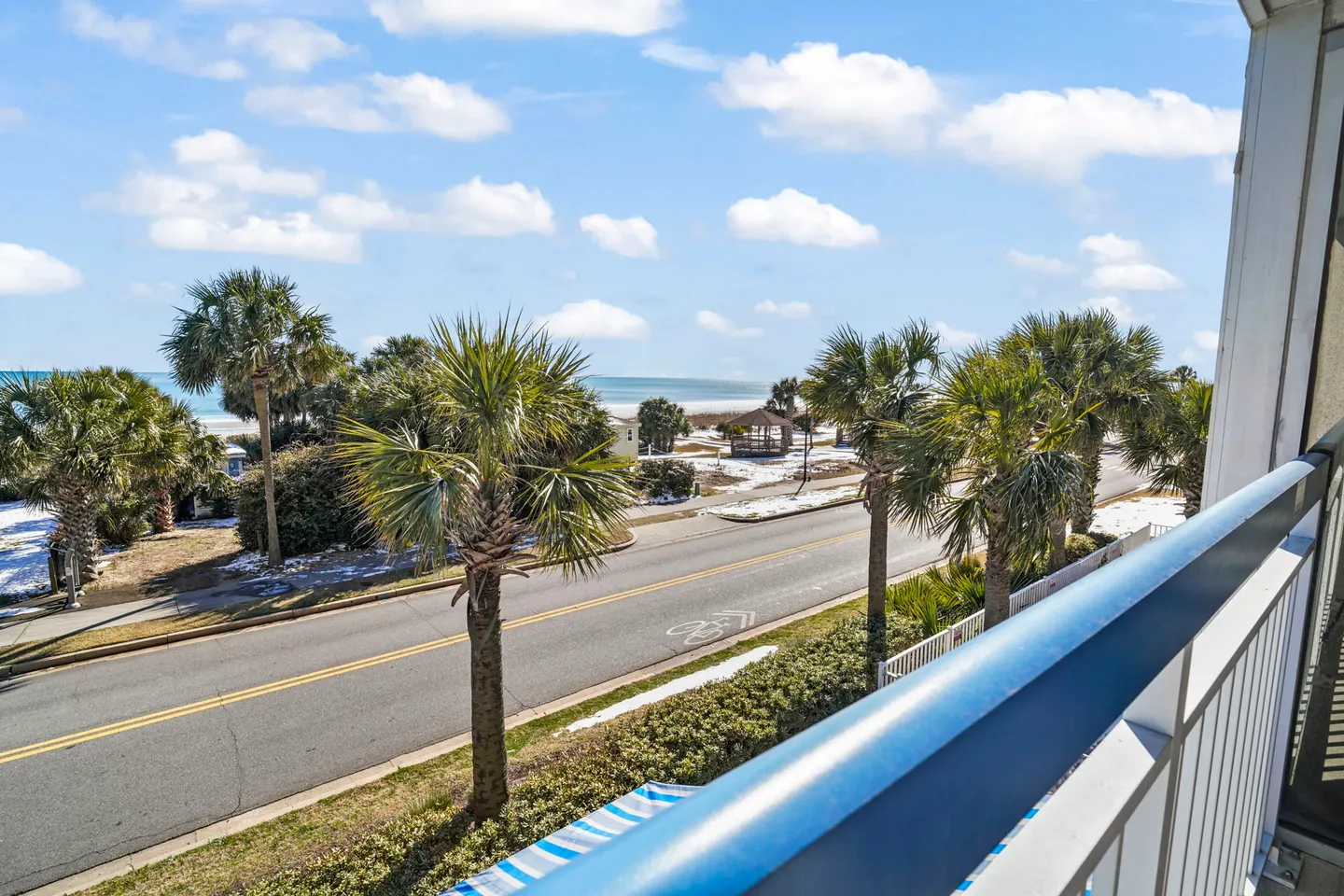 View from an elevated balcony looking across a coastal road lined with palm trees toward the ocean and beach under a bright blue sky.