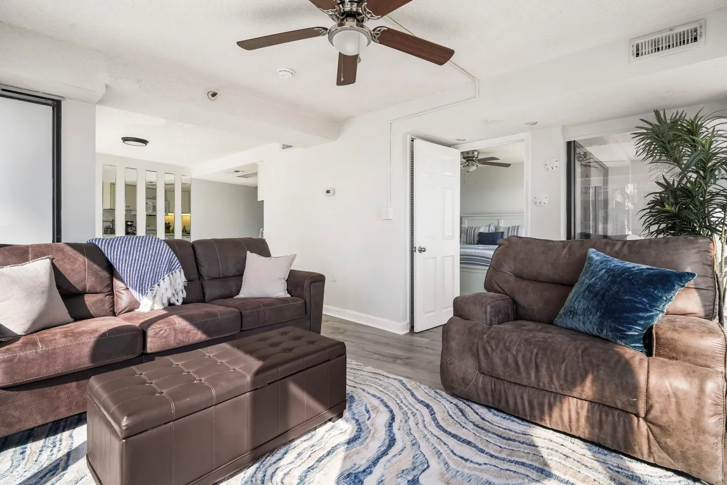 Living room with brown upholstered sofa, armchair, matching tufted ottoman, and a doorway opening to a bedroom.