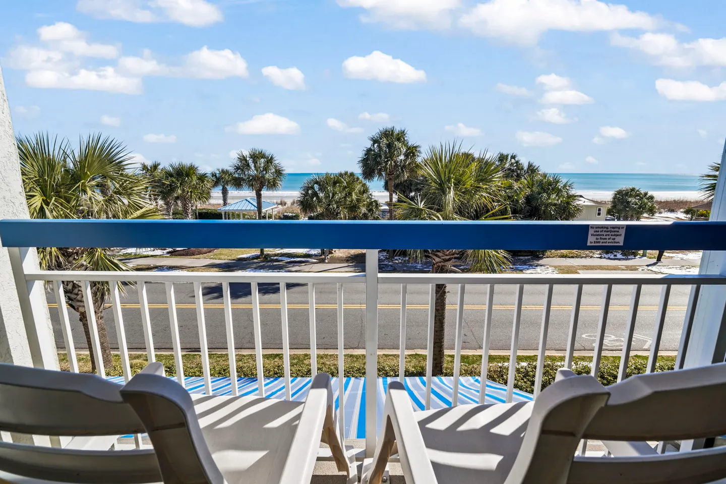 Balcony view overlooking palm trees, a road, and the ocean under a blue sky.