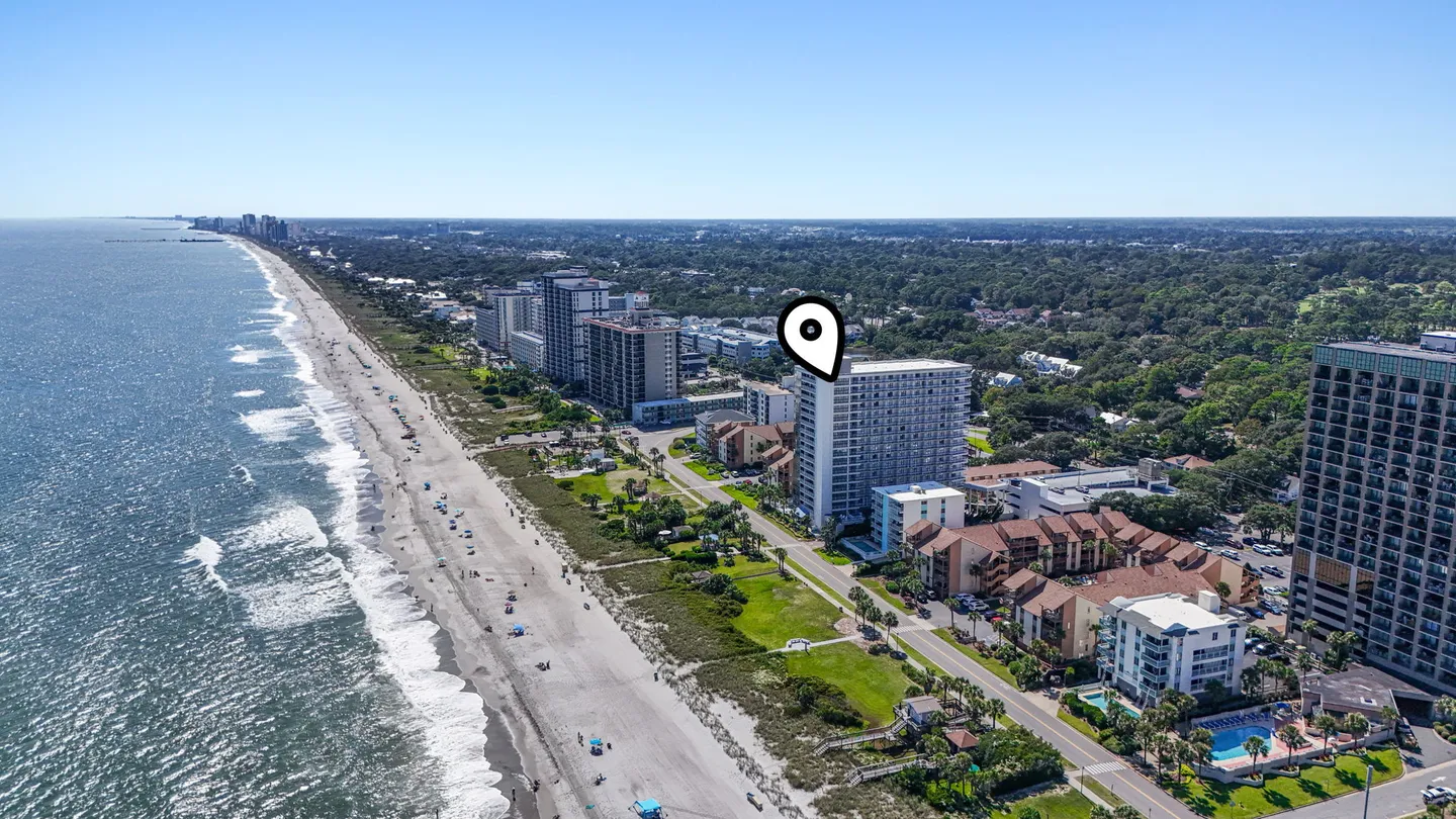 Aerial view of a coastal beach lined with multi-story buildings and ocean waves under a clear blue sky, marked with a location pin.