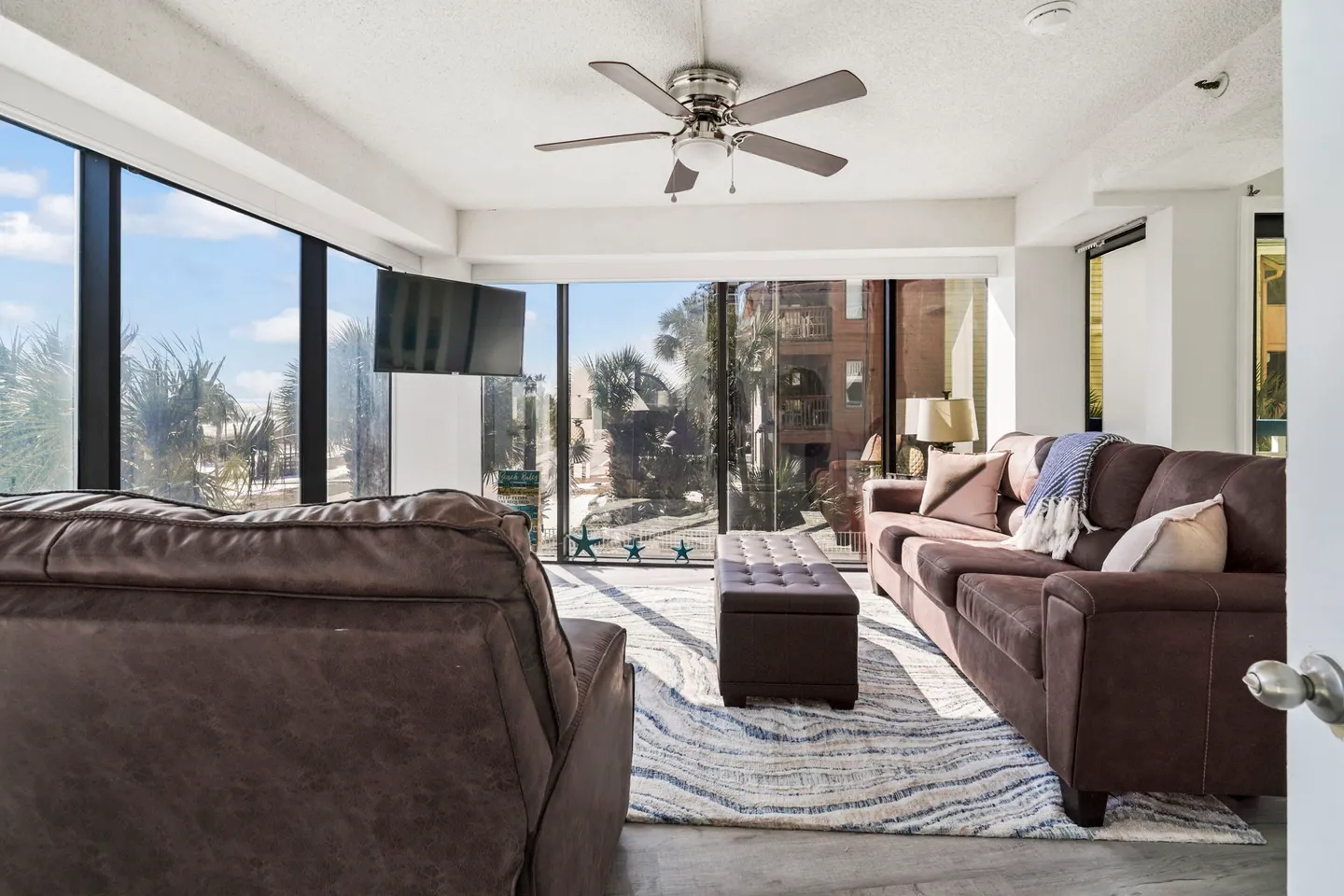 Sunlit living area with floor-to-ceiling windows, brown sofa, recliner, ottoman, and wall-mounted TV.