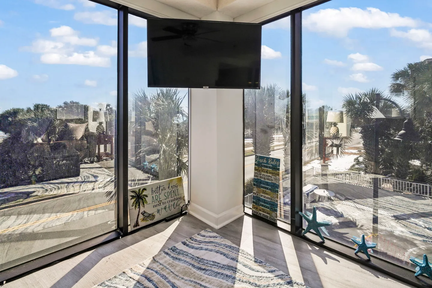 Corner window alcove with floor-to-ceiling glass panels framing views of outdoor palm trees and blue sky, with a mounted television overhead.