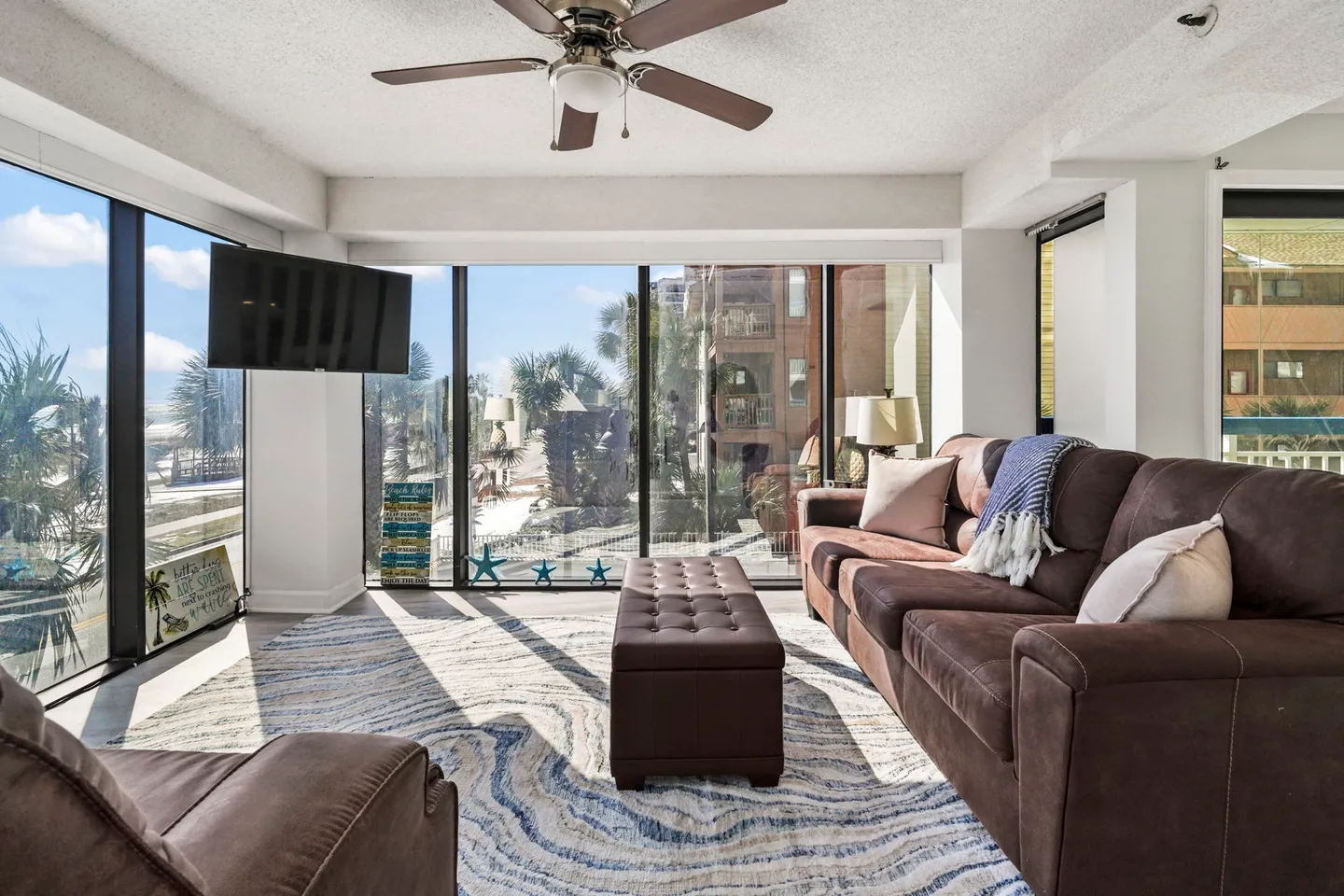 Sunlit living room with large glass windows, a brown sectional sofa, matching ottoman, ceiling fan, and wall-mounted TV.