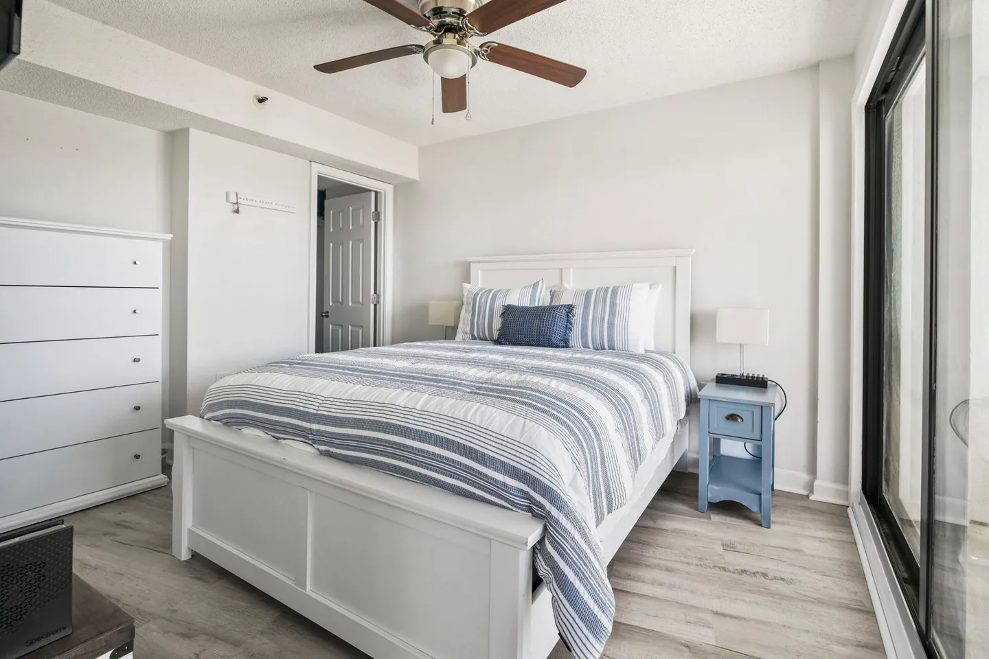 Bedroom with a white panel bed dressed in blue-and-white striped bedding, flanked by a blue nightstand and a tall white dresser next to sliding glass doors.