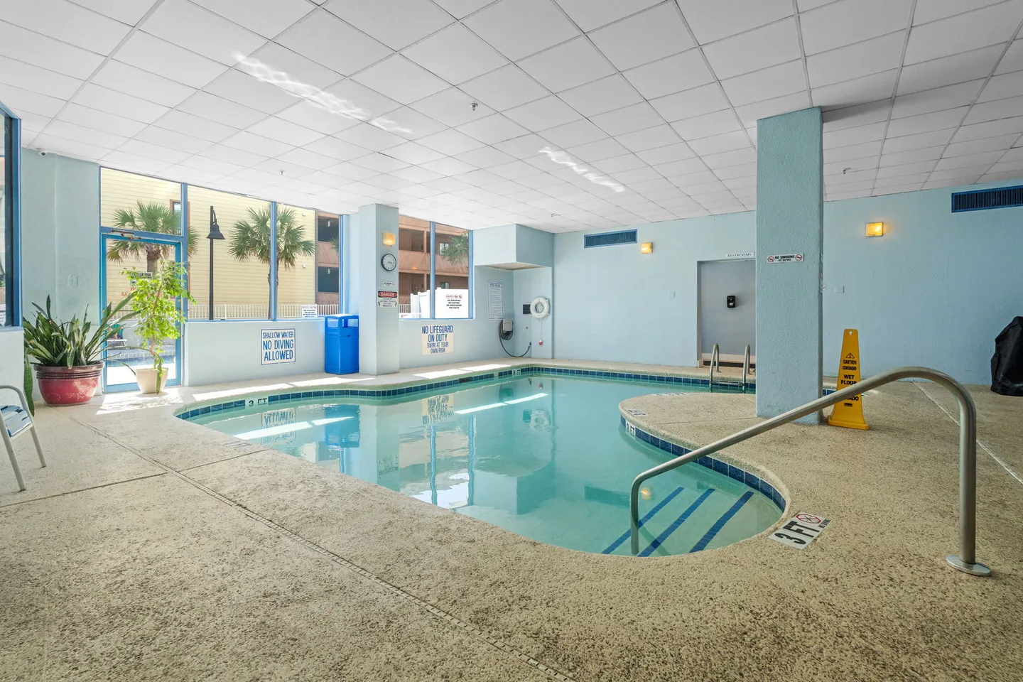 Indoor swimming pool with light blue walls, tiled ceiling, and large windows looking outside.