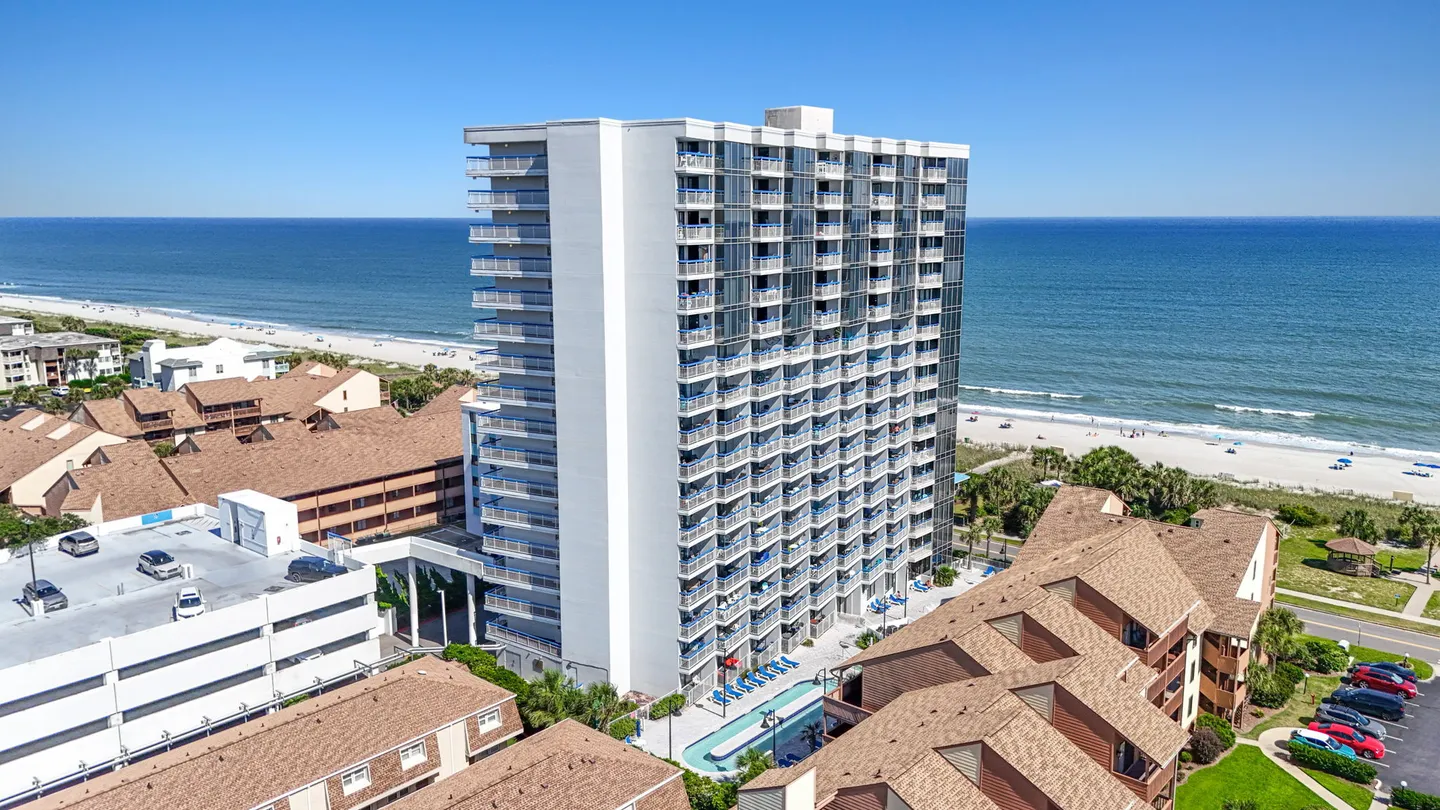 Aerial view of a multistory white high-rise beachfront condominium building next to the ocean and sandy beach.