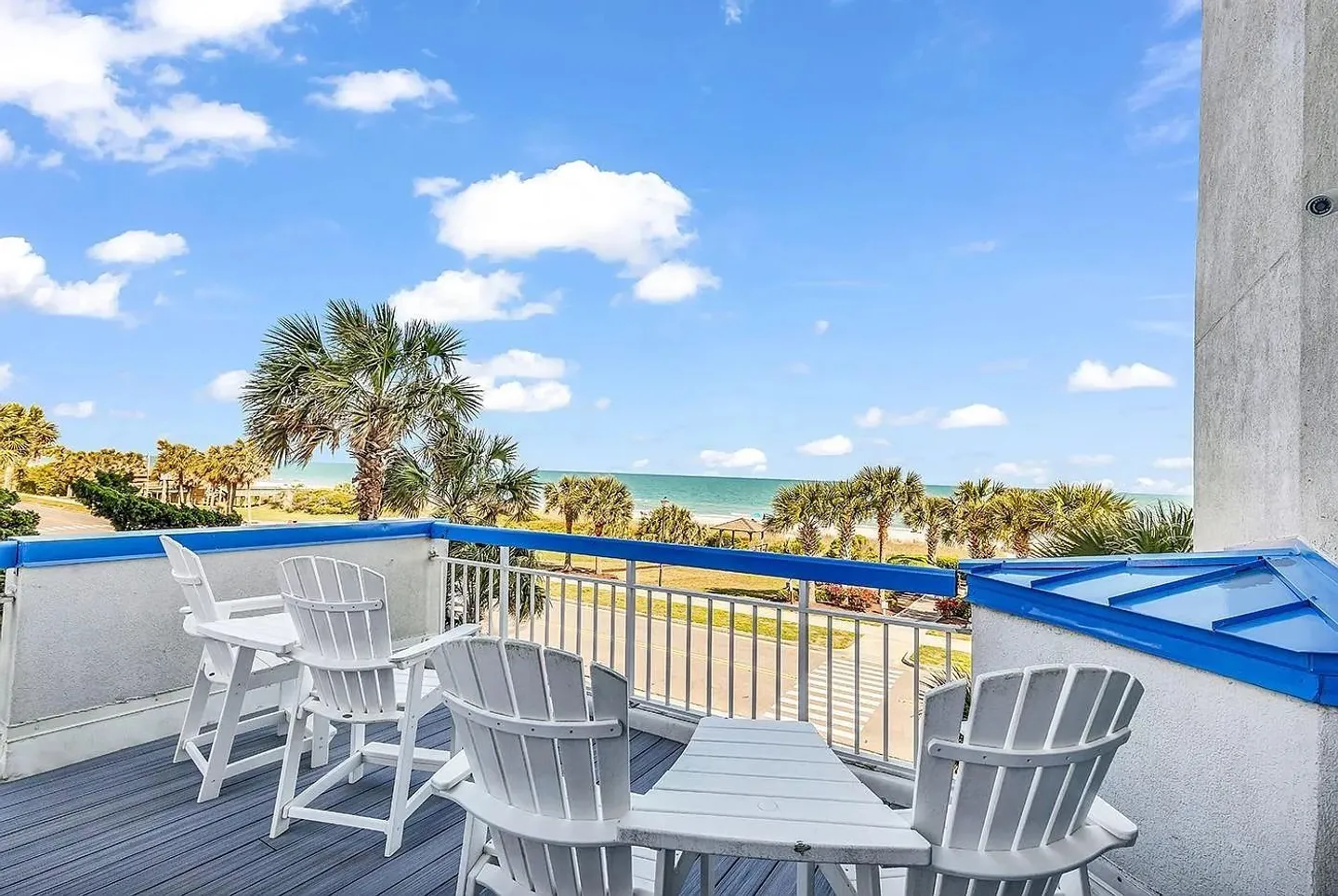 Balcony with white chairs and small tables overlooking palm trees and the ocean under a blue sky.