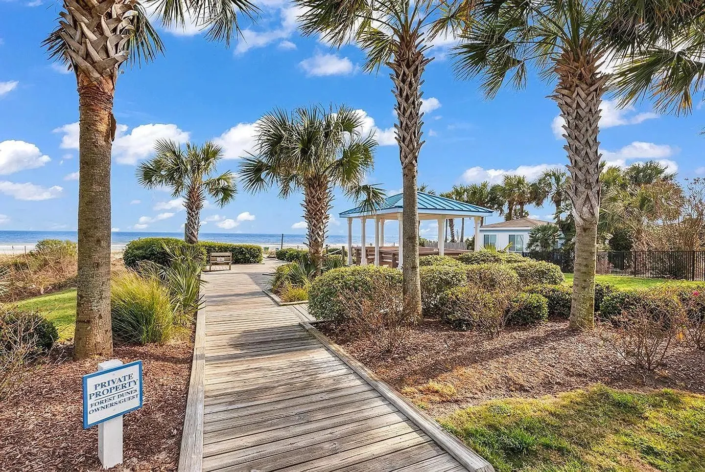 A wooden boardwalk pathway lined with palm trees leading toward the ocean and beach, with a gazebo on the right under a blue sky.