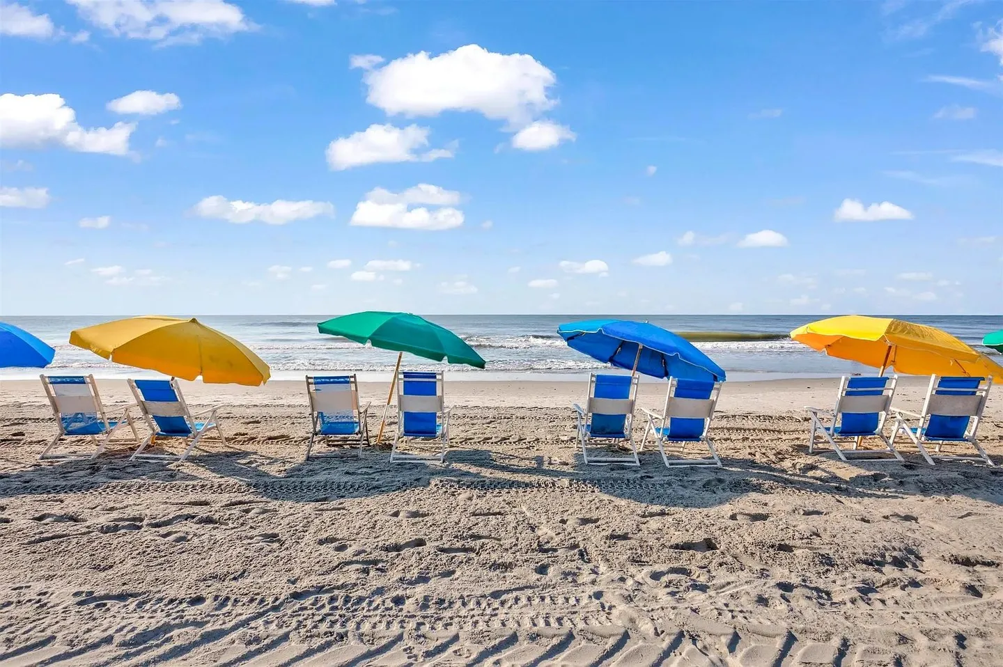 Line of beach chairs with colorful umbrellas set up on the sand facing the ocean beneath a sunny sky with clouds.