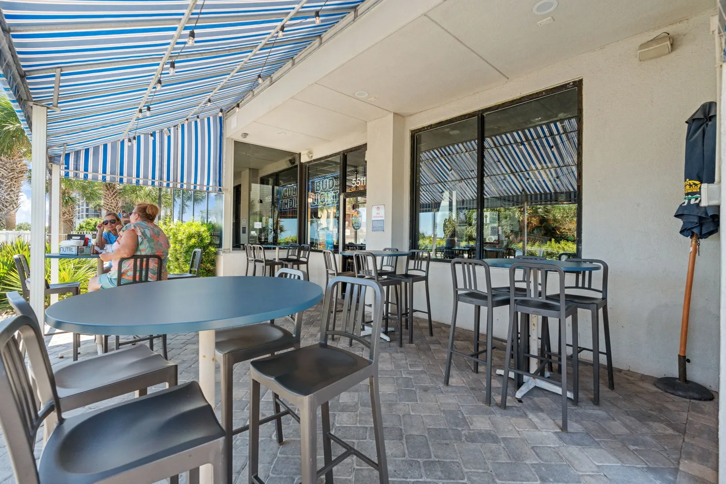 Outdoor patio dining area with paved stone ground, high-top tables, chairs, and a blue and white striped awning.