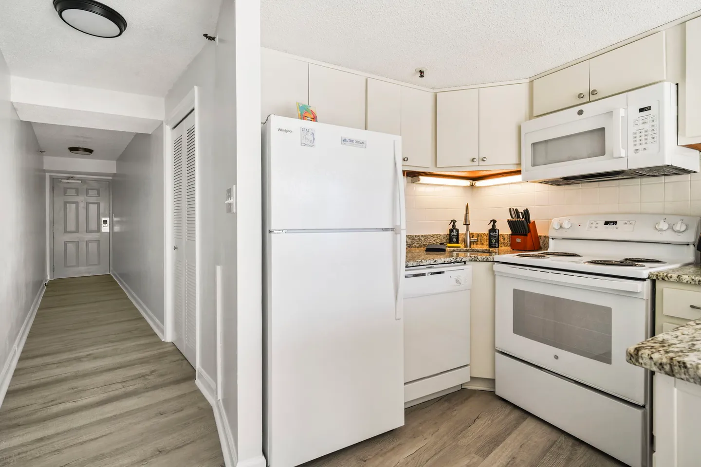 Kitchen with white appliances, light-colored cabinets, and speckled countertops next to an entryway hallway.