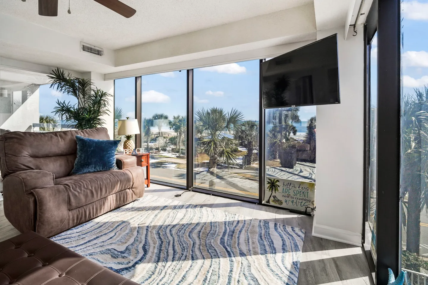Living area with plush seating, patterned area rug, mounted television, and large floor-to-ceiling windows showing palm trees and the beach.