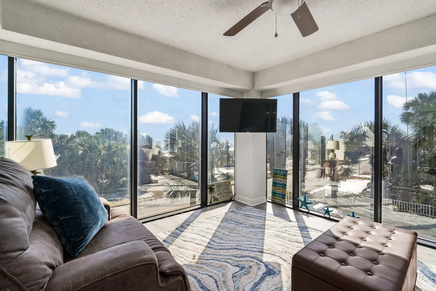 Sunroom seating area featuring panoramic floor-to-ceiling windows with palm tree views, an armchair, ottoman, and wall-mounted television.