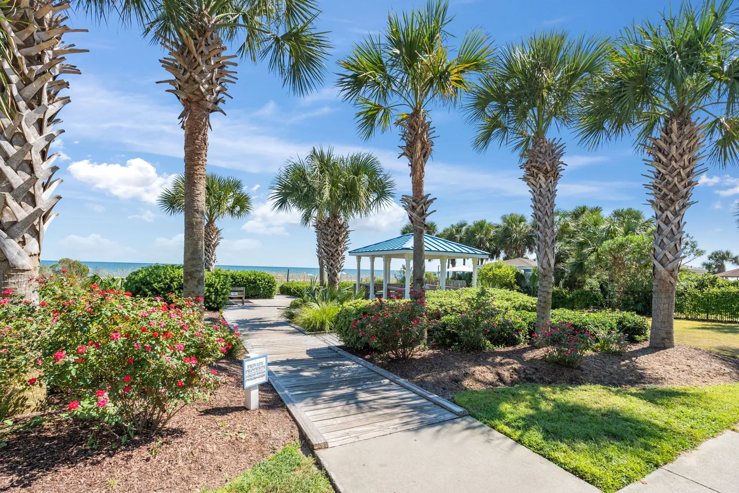 Wooden boardwalk flanked by palm trees and flowering bushes leading toward a beachfront gazebo and ocean.