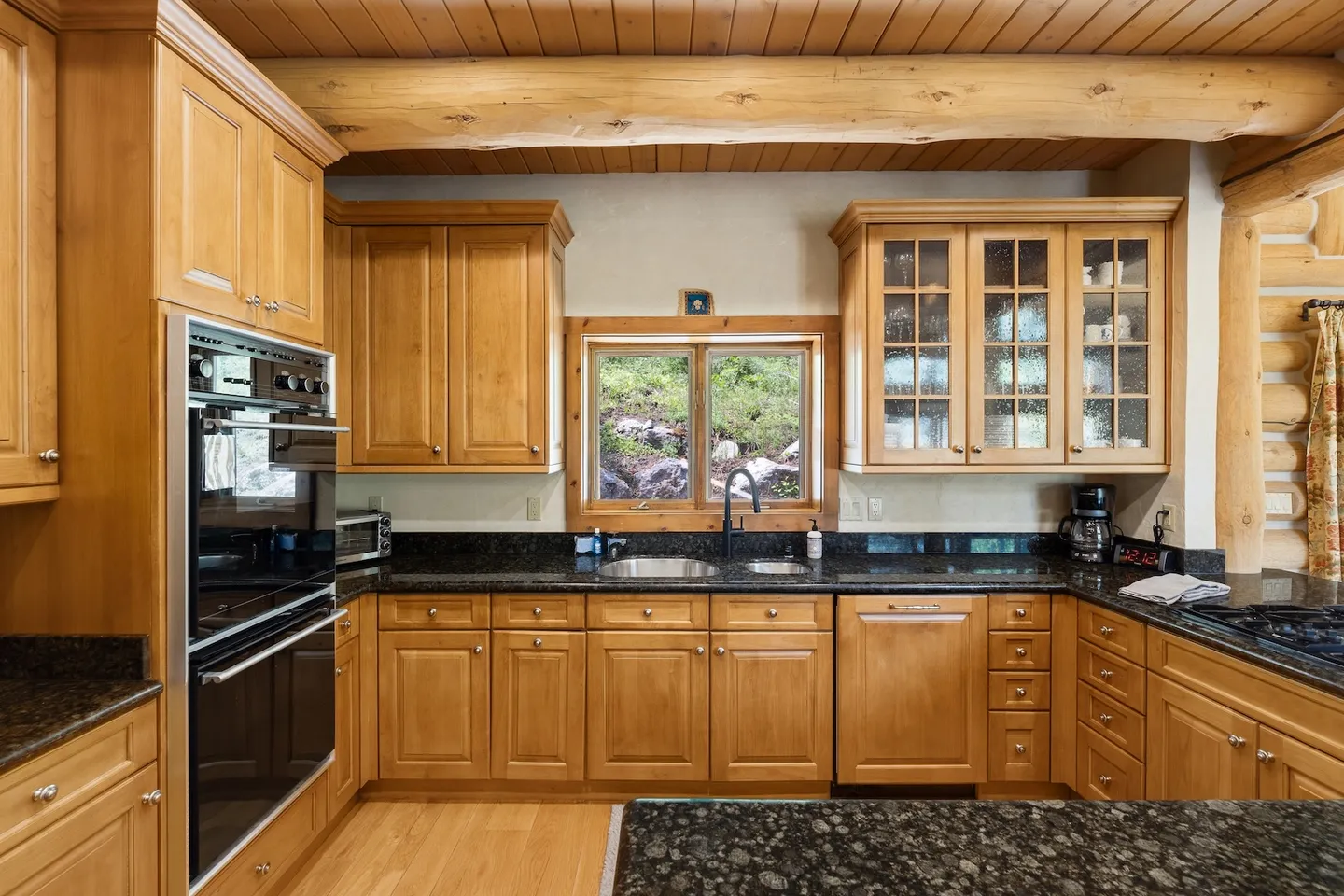 Kitchen with light wood cabinetry, dark granite countertops, built-in double oven, and exposed log ceiling beams.