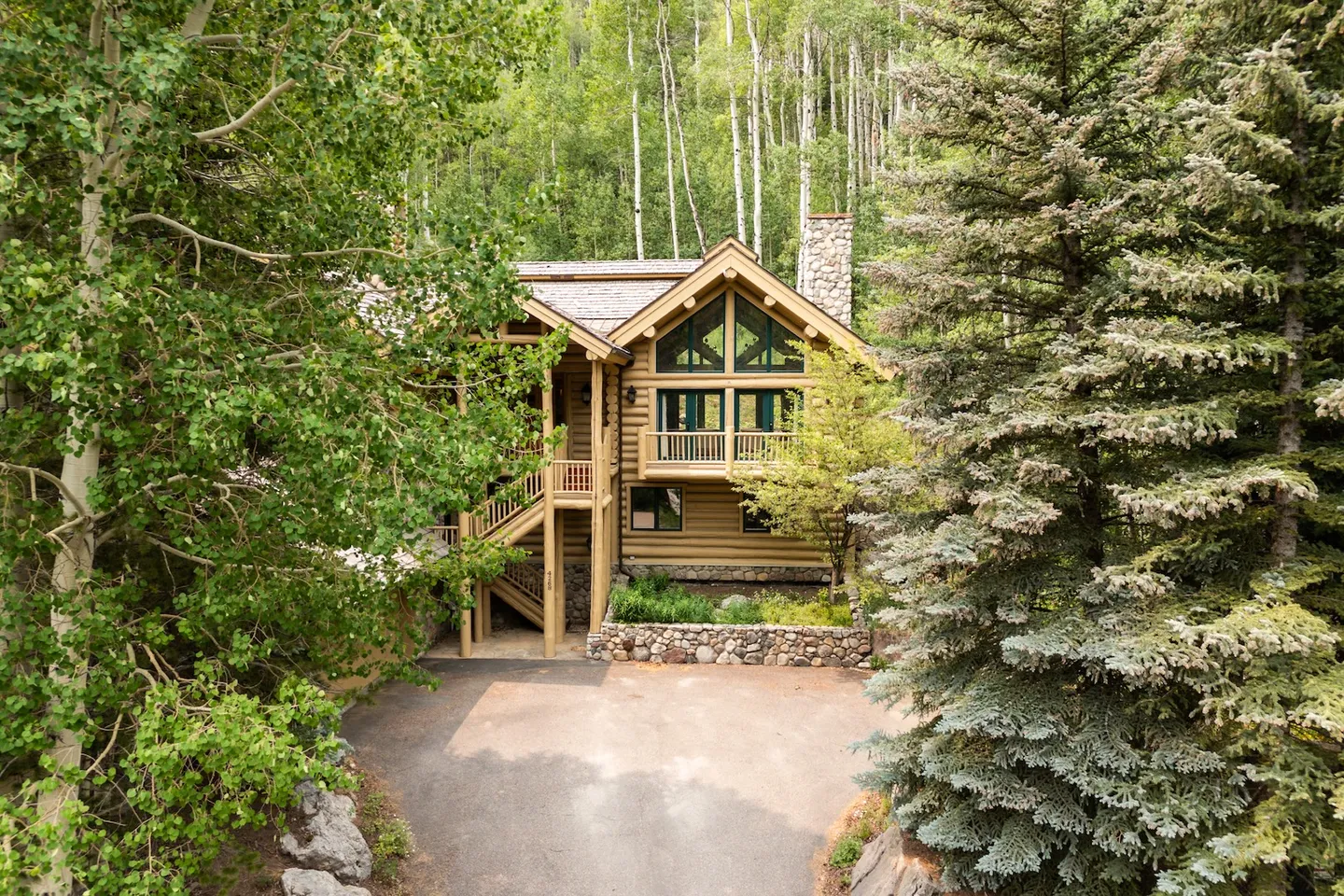 Exterior view of a multi-story log cabin style home surrounded by dense pine and aspen trees with a paved driveway in the foreground.